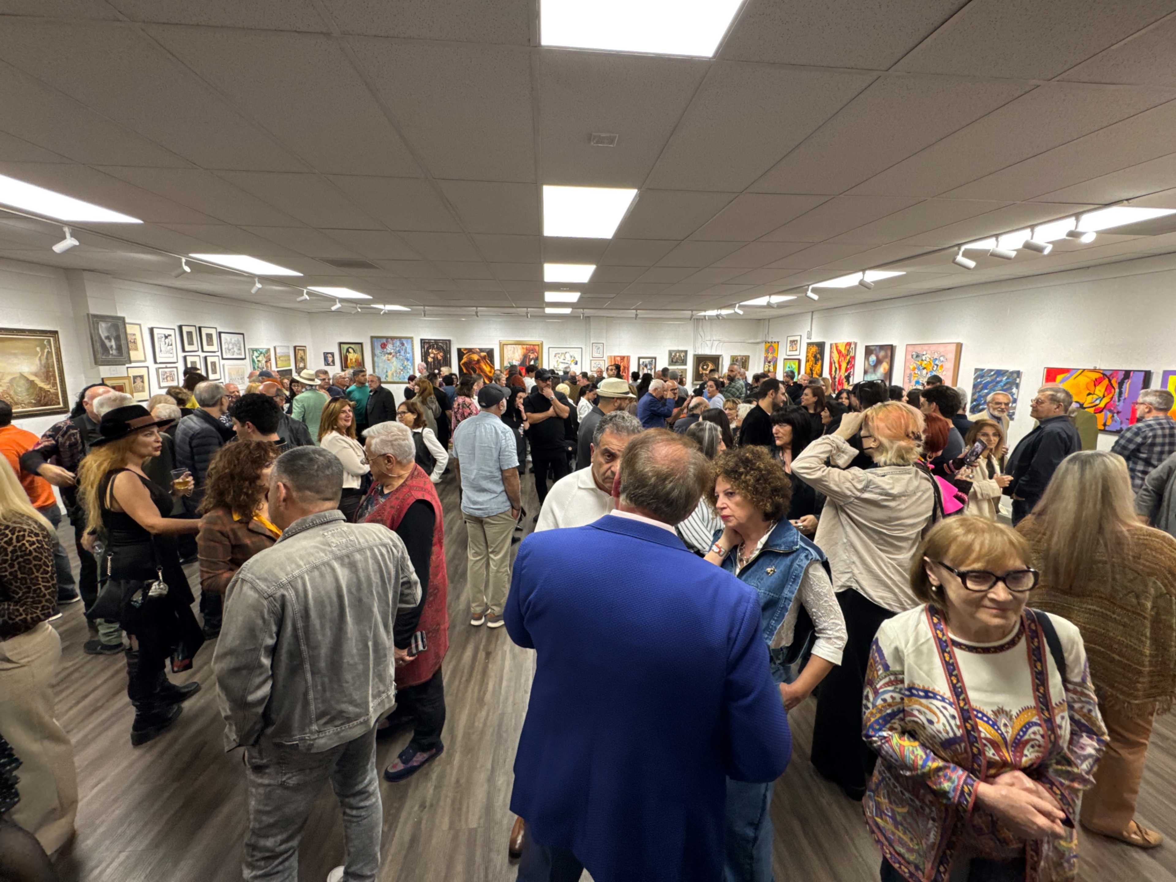 A large crowd of people is gathered in an art gallery, surrounded by various artworks displayed on the walls.