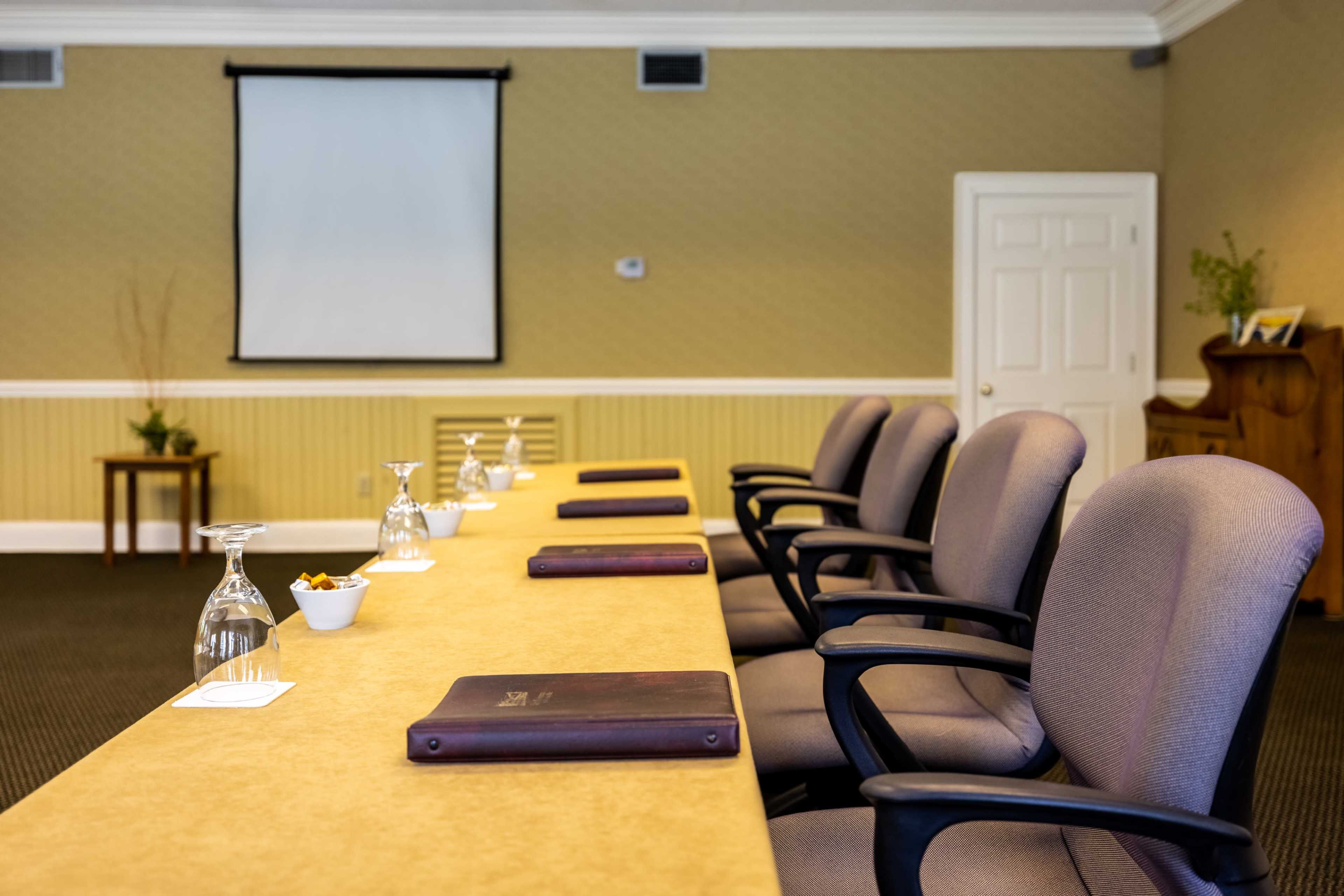 A conference room features a long table with glasses, notepads, and a projector screen in the background.