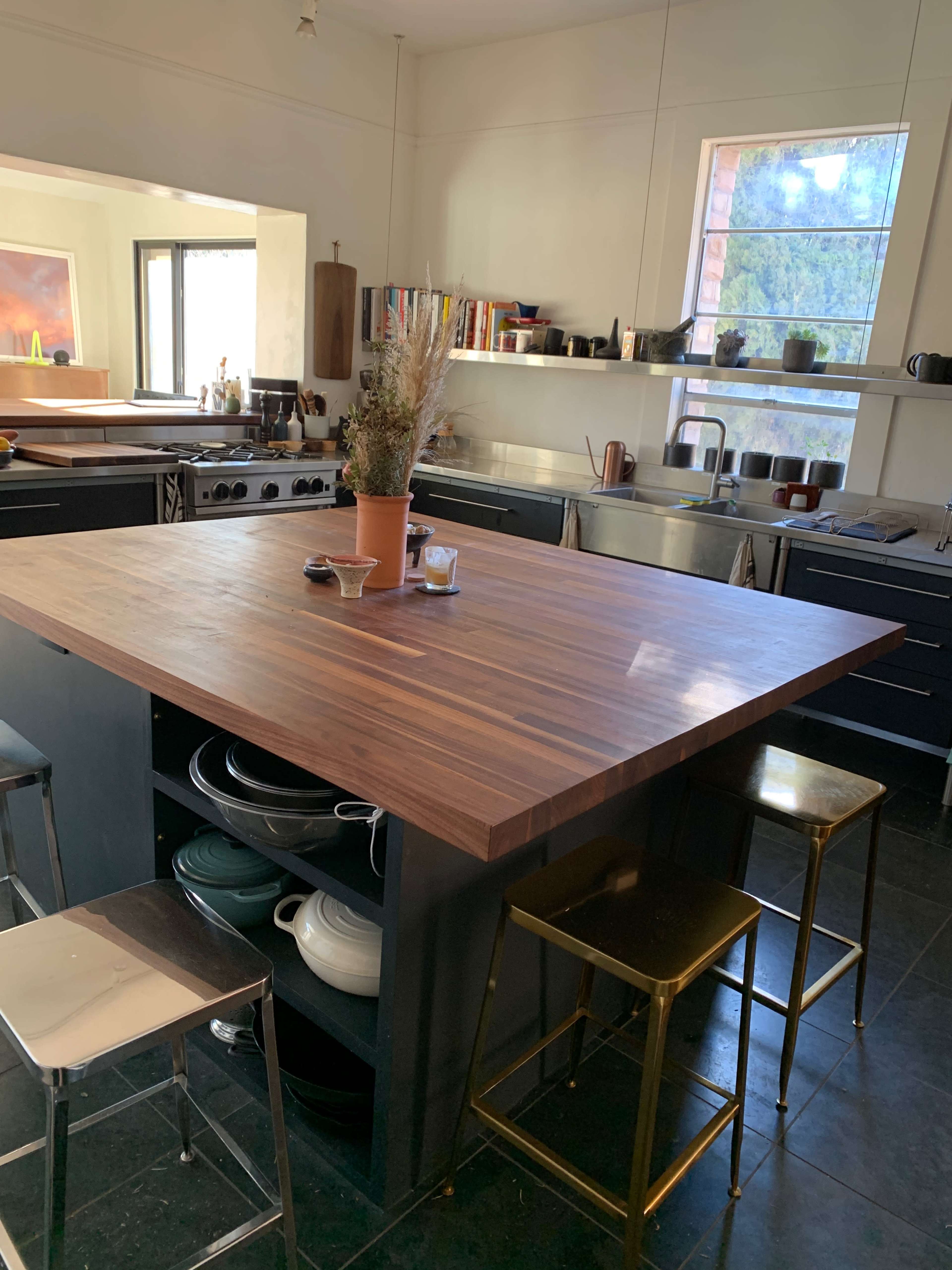 The image shows a modern kitchen with a large wooden island, surrounded by metal stools, and shelves filled with various kitchenware.