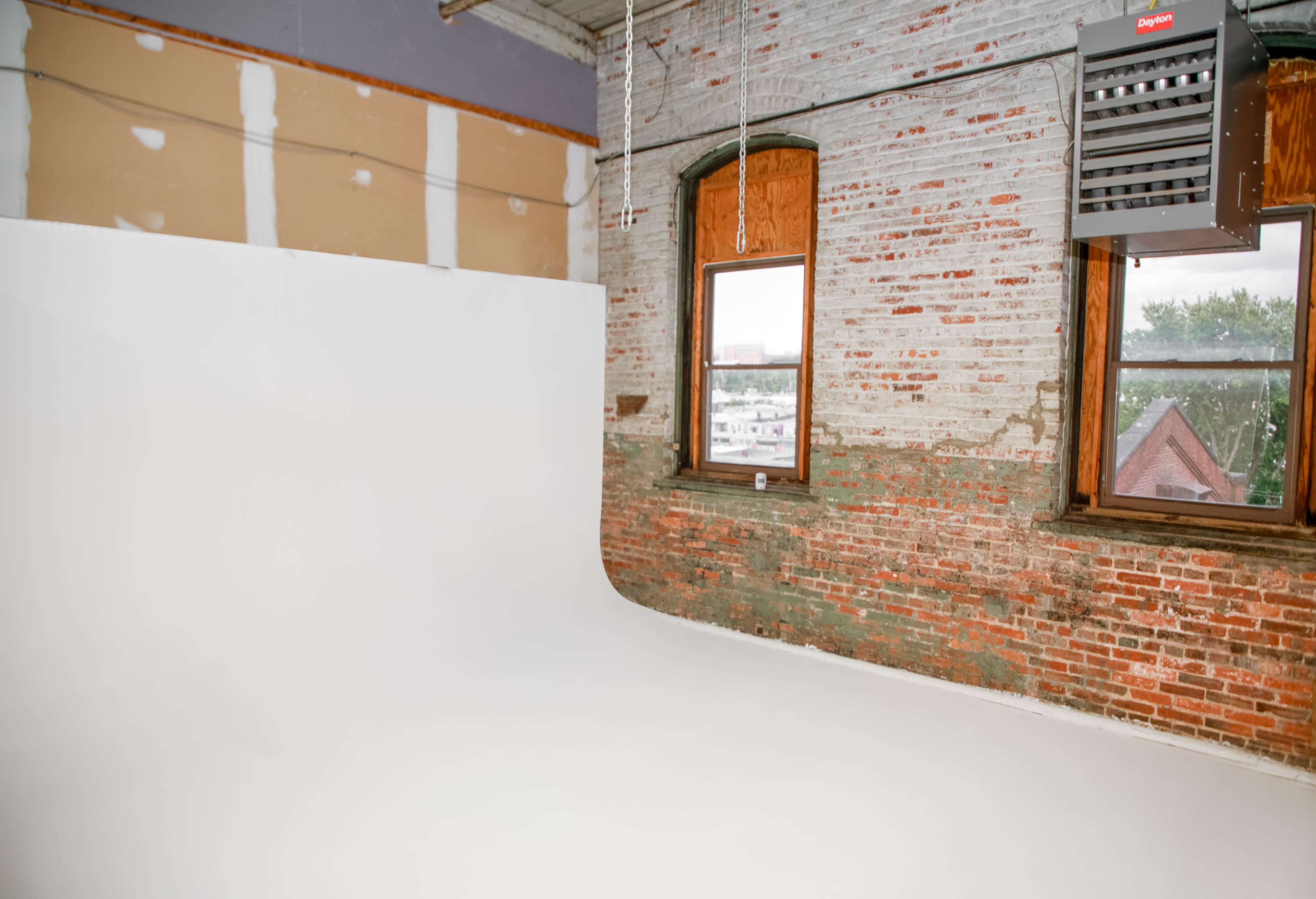 A large, empty photography studio features a seamless white backdrop and exposed brick walls with a window revealing a view outside.