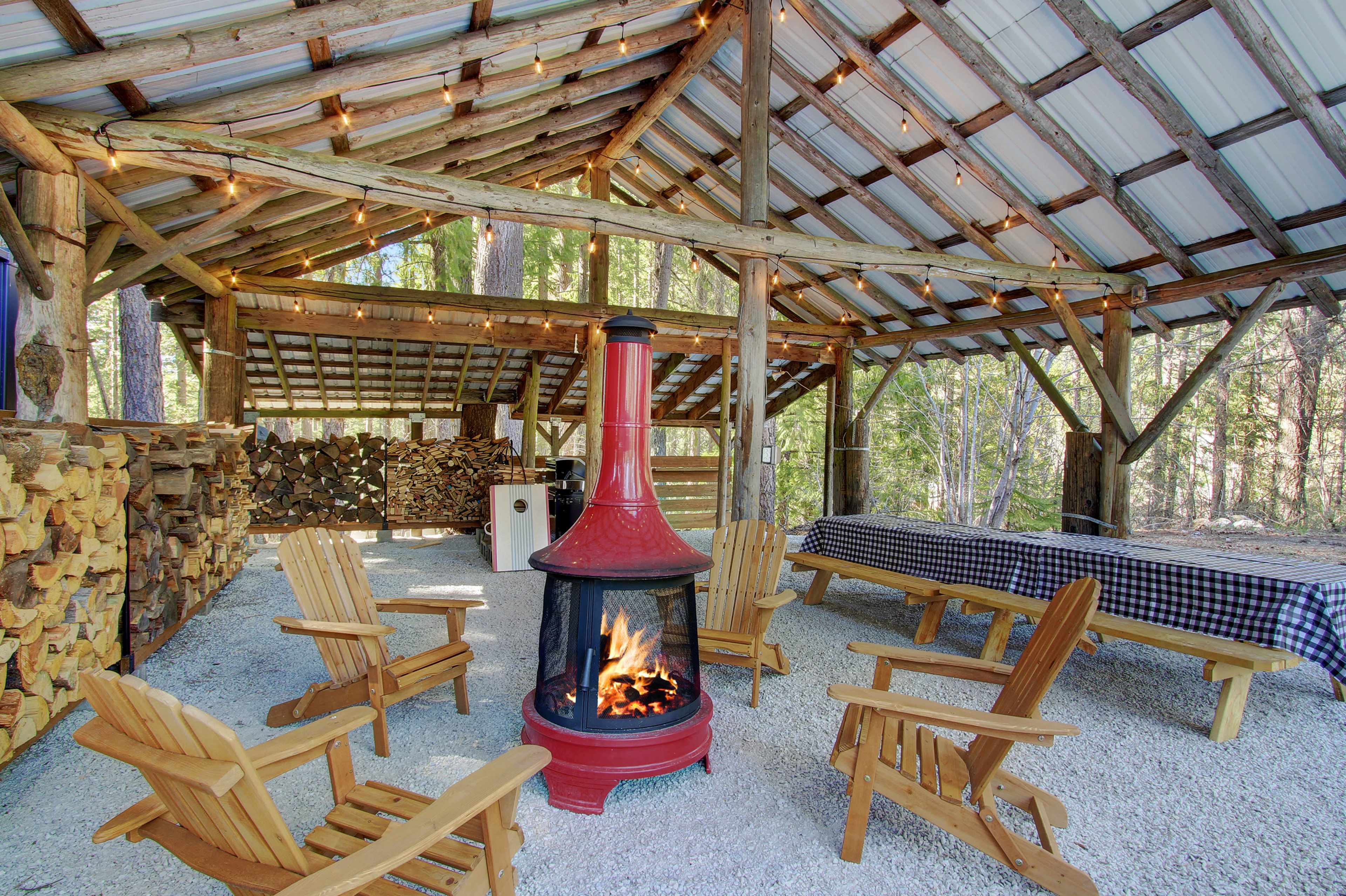 The image shows a rustic outdoor shelter featuring a red wood stove surrounded by wooden chairs, a long table, and stacked firewood in the background.