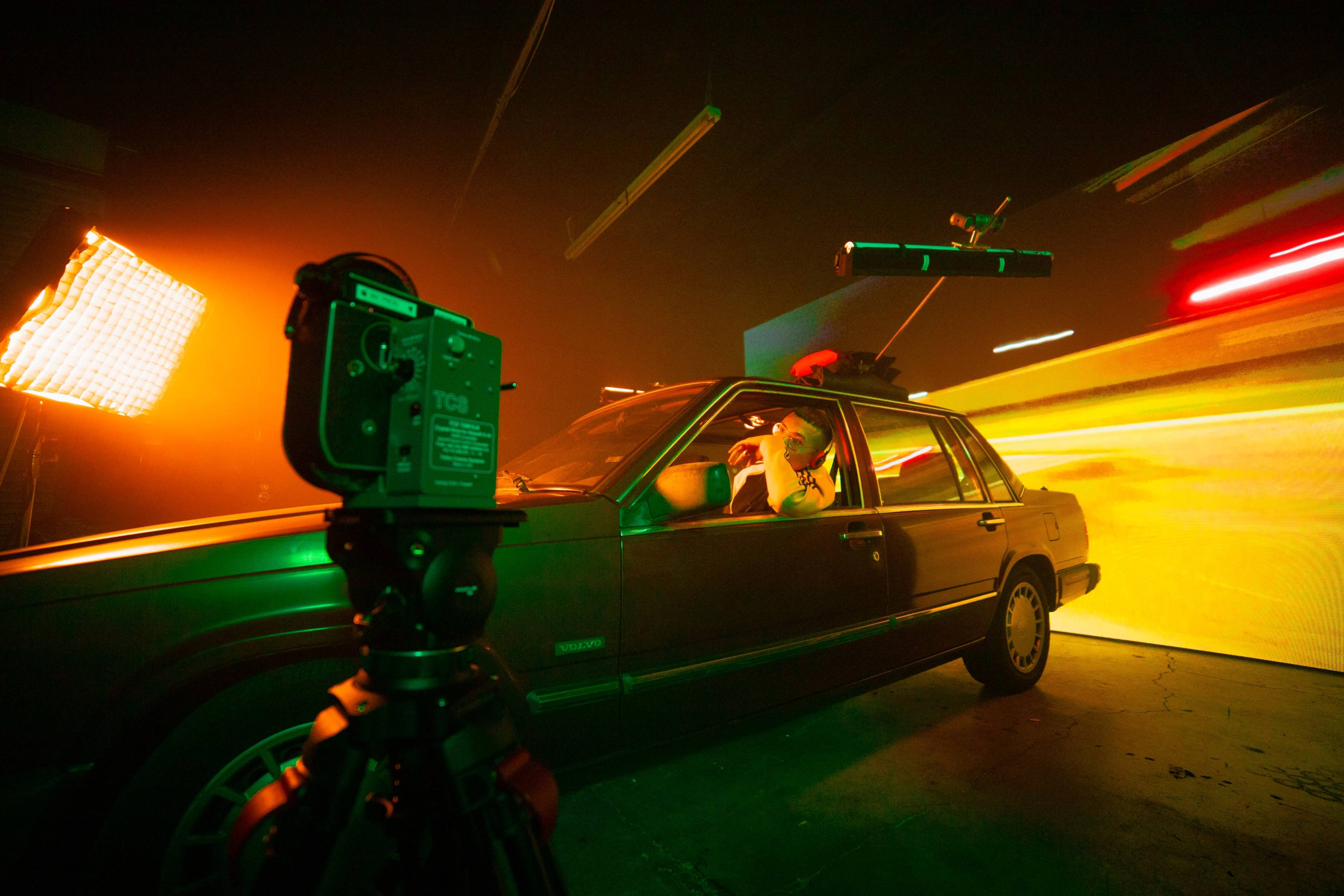 A person sits inside a car while a camera on a tripod captures the scene against a backdrop of colorful lighting effects.