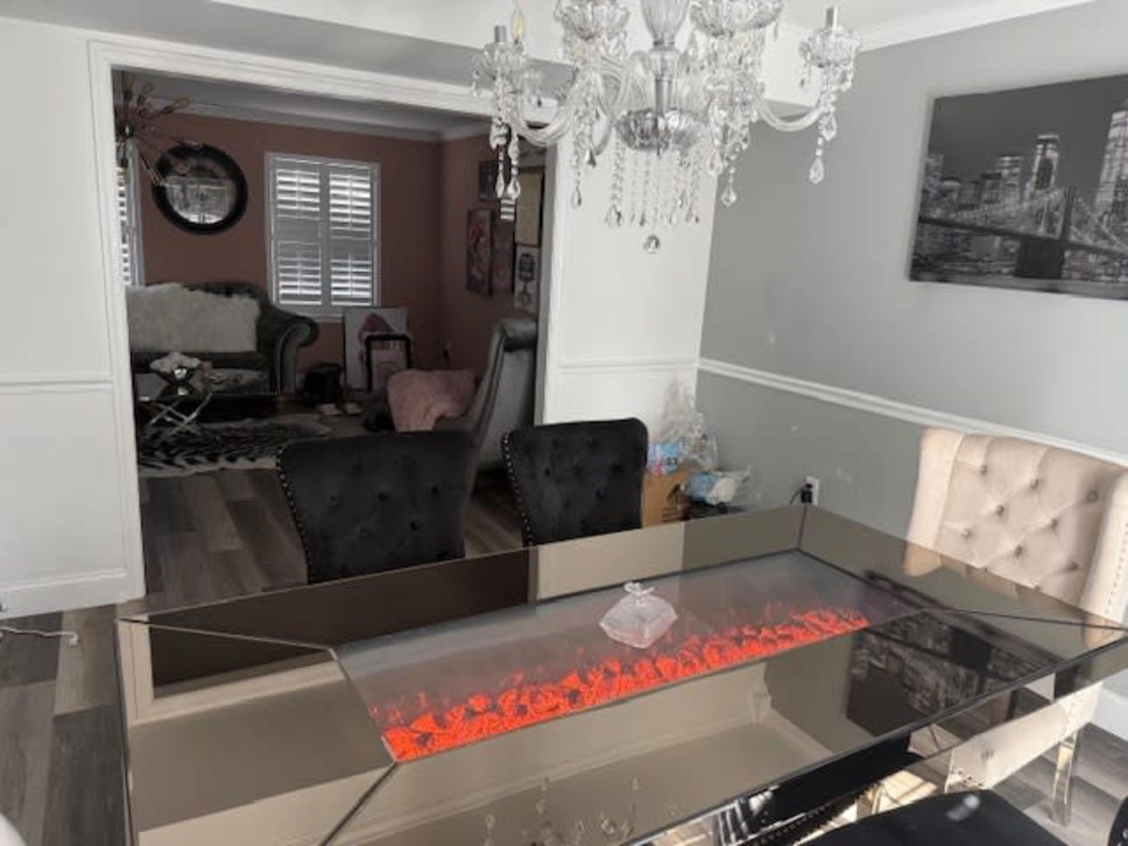 The image shows a modern dining room featuring a glass table with a built-in flame light effect and plush tufted chairs, with an adjacent living area visible.