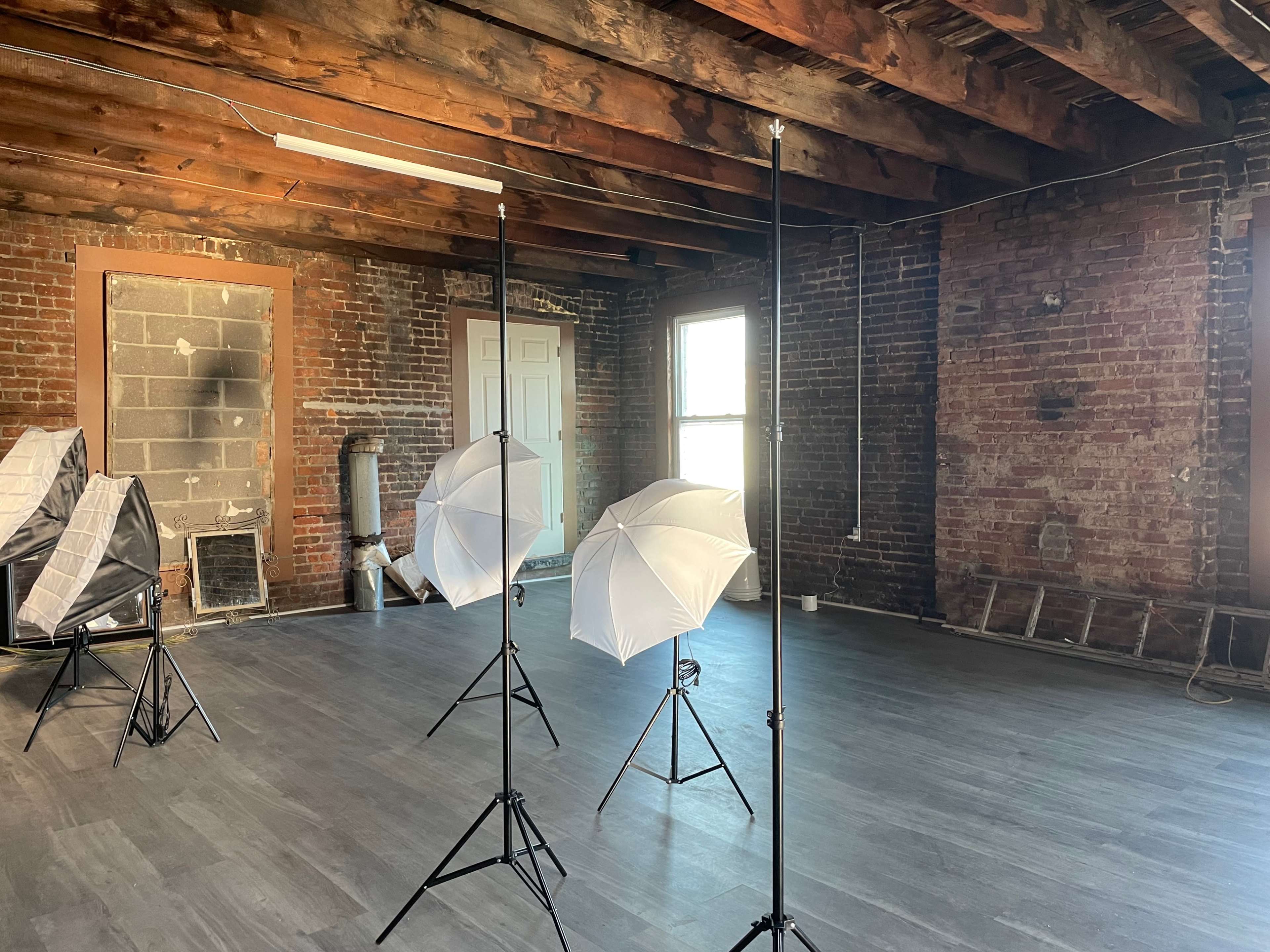 An empty room with exposed brick walls, wooden beams, and three photo lighting setups with white umbrellas.