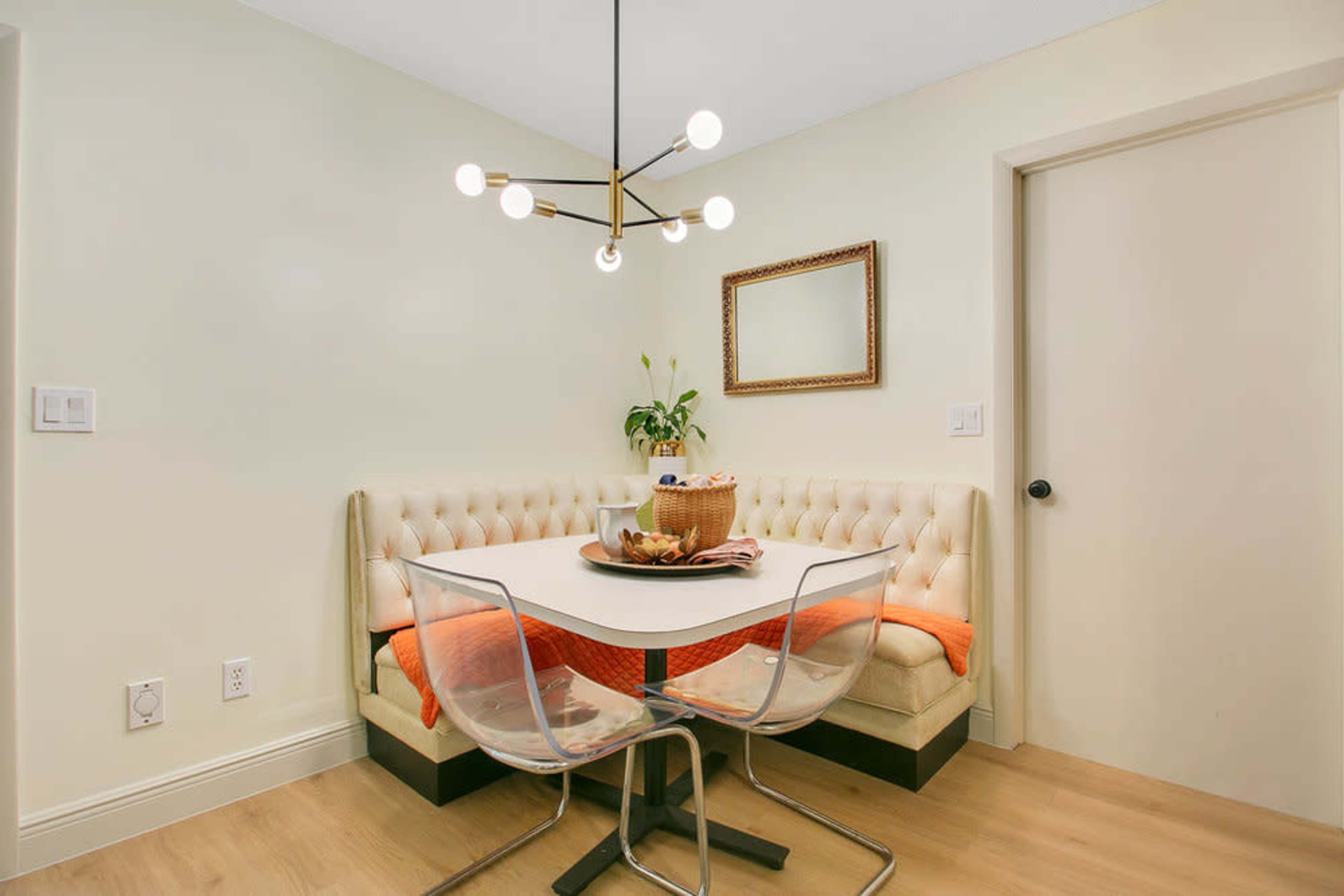 A small dining area features a beige tufted banquette, a round white table, clear acrylic chairs, and decorative items, including a bowl of fruit on the table.