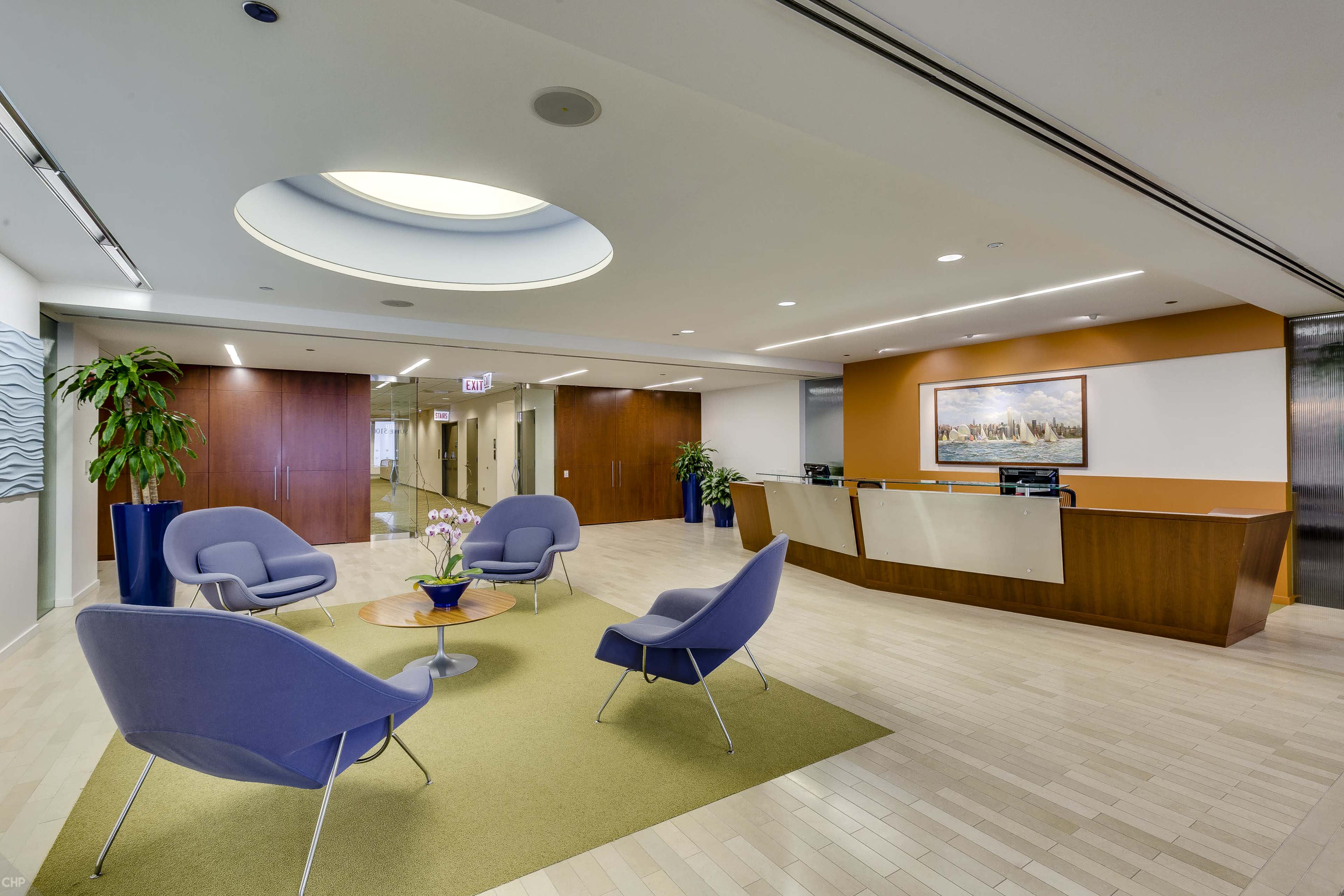A modern office lobby with a reception area, four purple chairs arranged around a circular green rug, and a large circular skylight above.