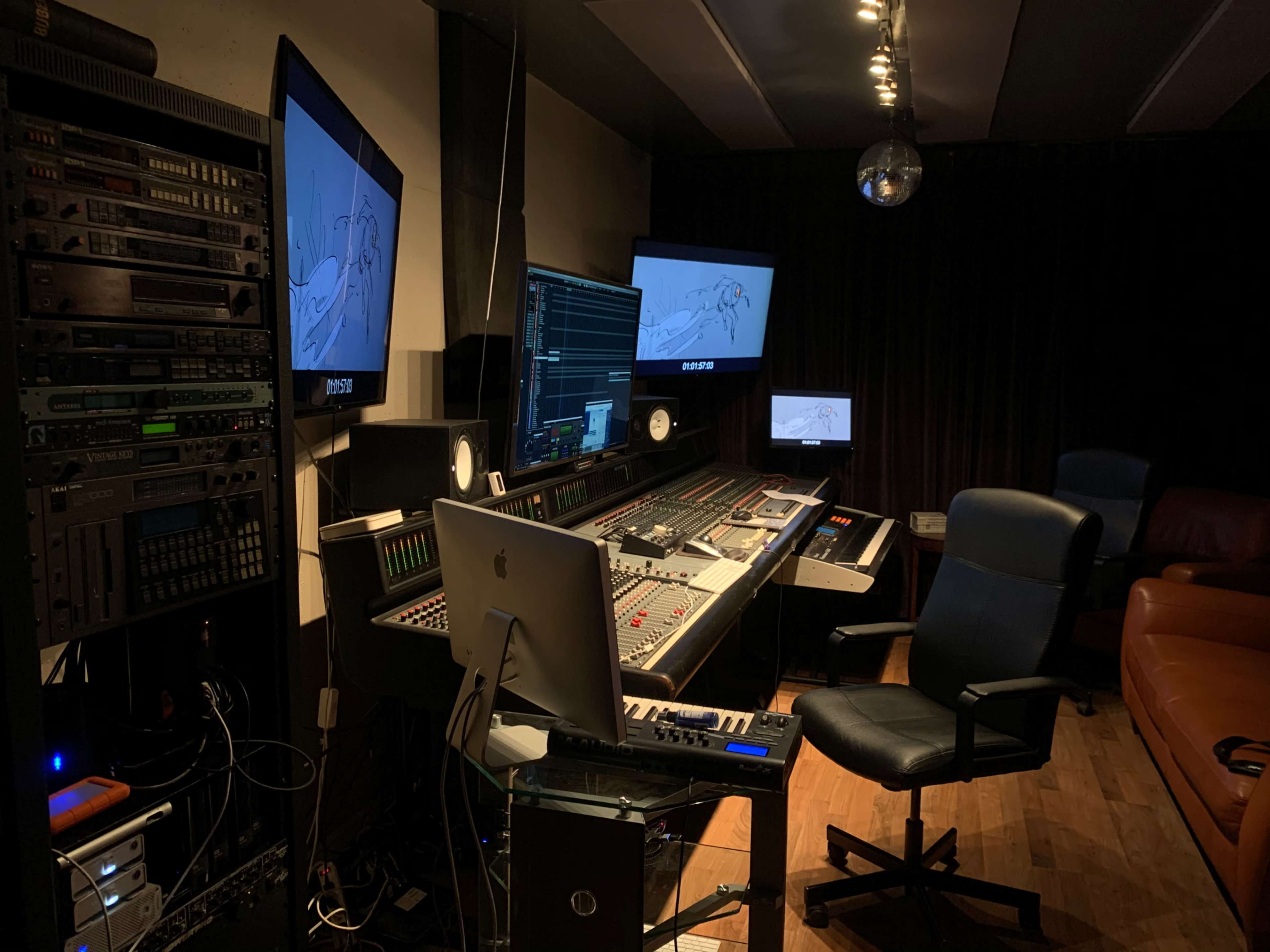 The image shows a music production studio equipped with a mixing console, two large monitors displaying audio waveforms, and various audio equipment on the walls.