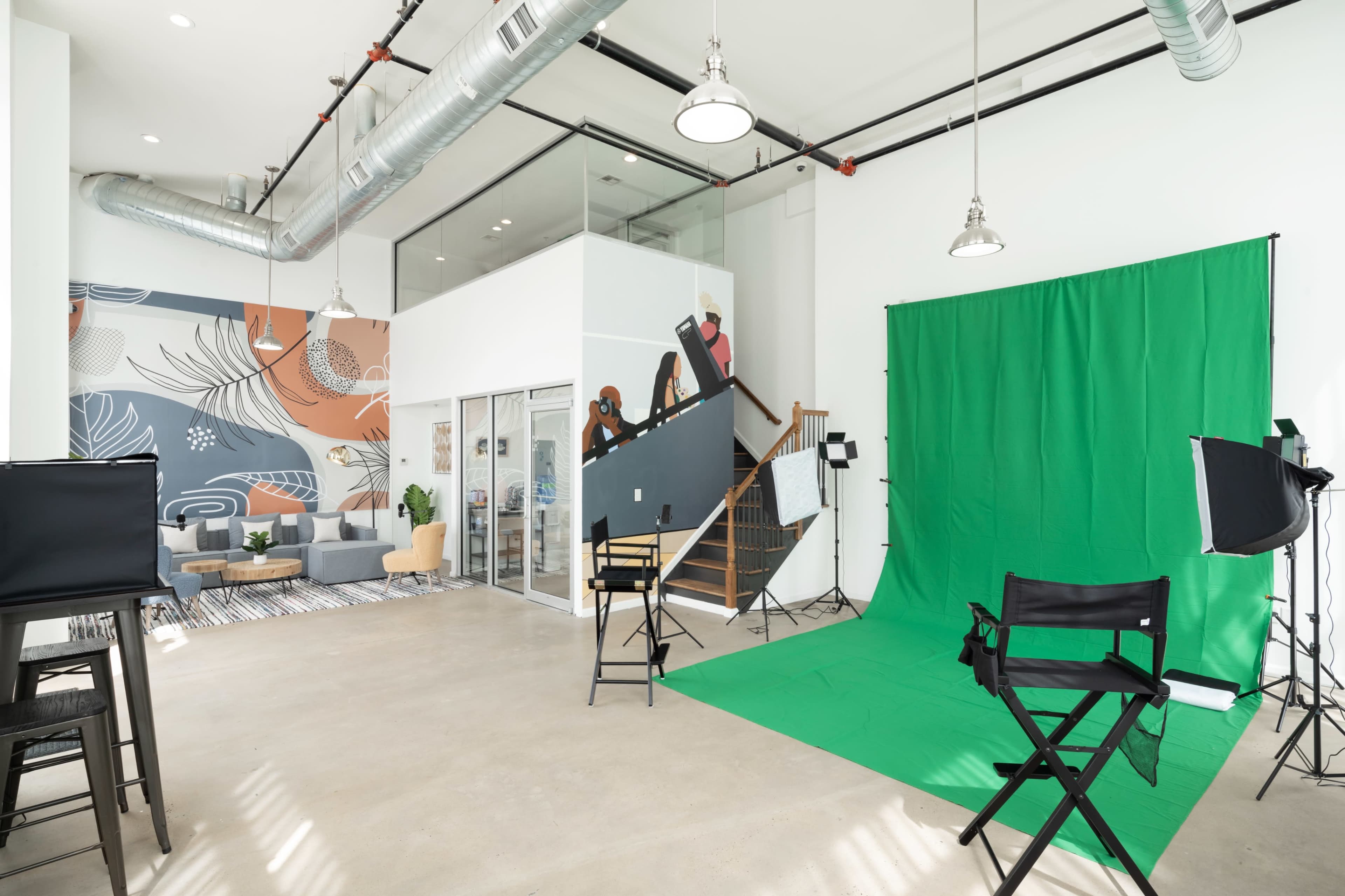 A modern studio space featuring a green screen setup, seating area, and a staircase, with industrial elements like exposed ductwork and large windows.