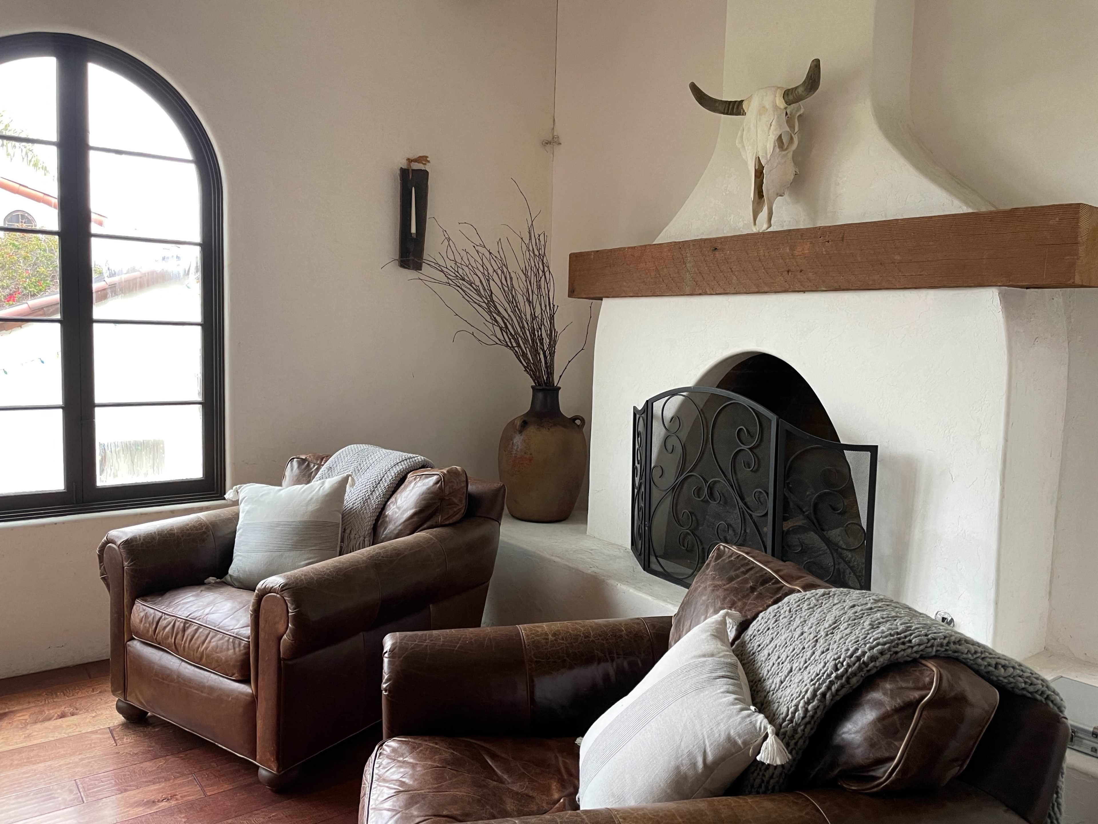 A living room with two brown leather armchairs, a rustic fireplace with a wooden mantel, and a decorative vase with branches placed beside the fireplace.