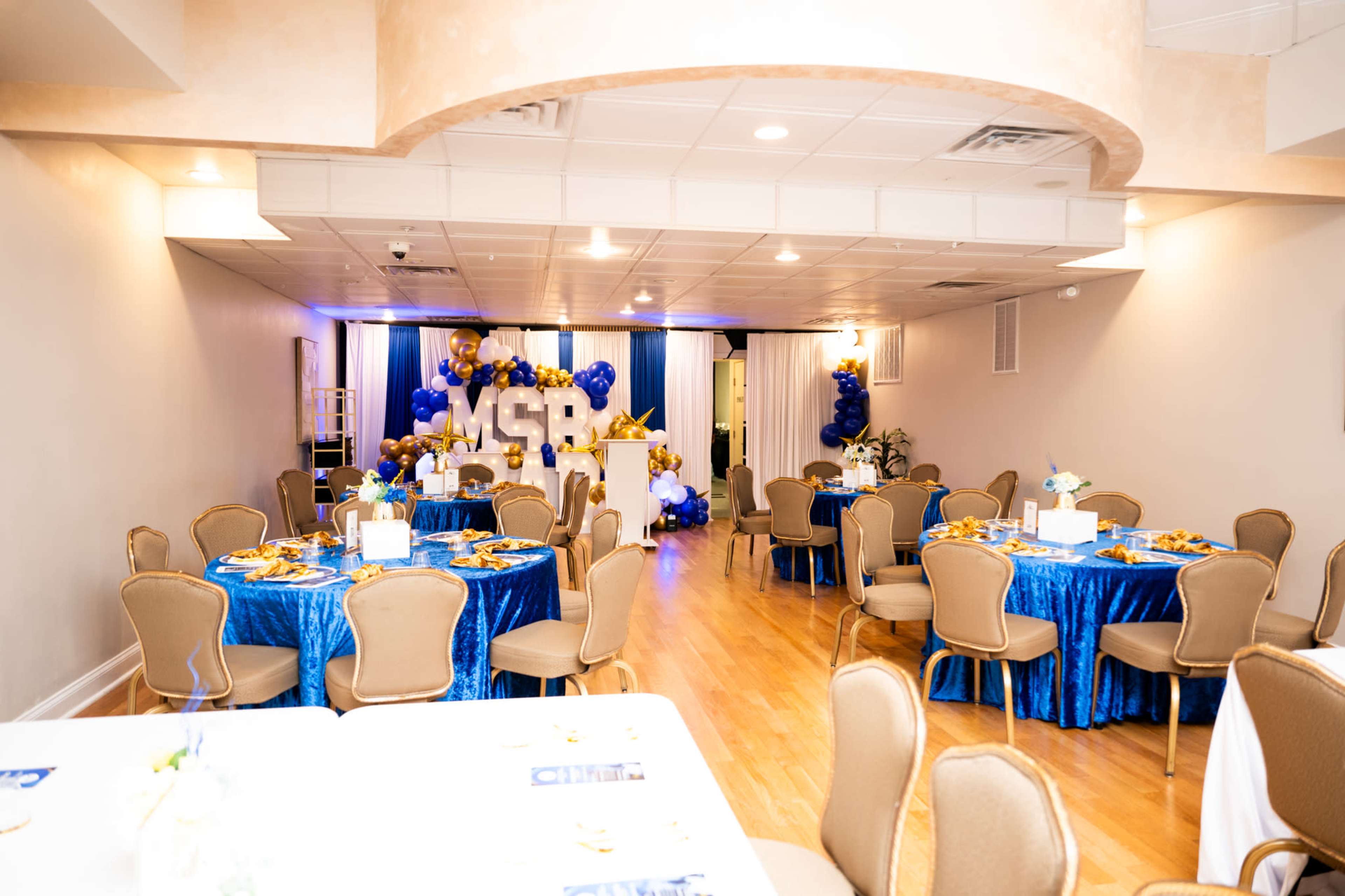 A banquet hall is decorated with blue and gold table settings, featuring round tables with white centerpieces, and a backdrop of balloons and letters spelling "MSR."