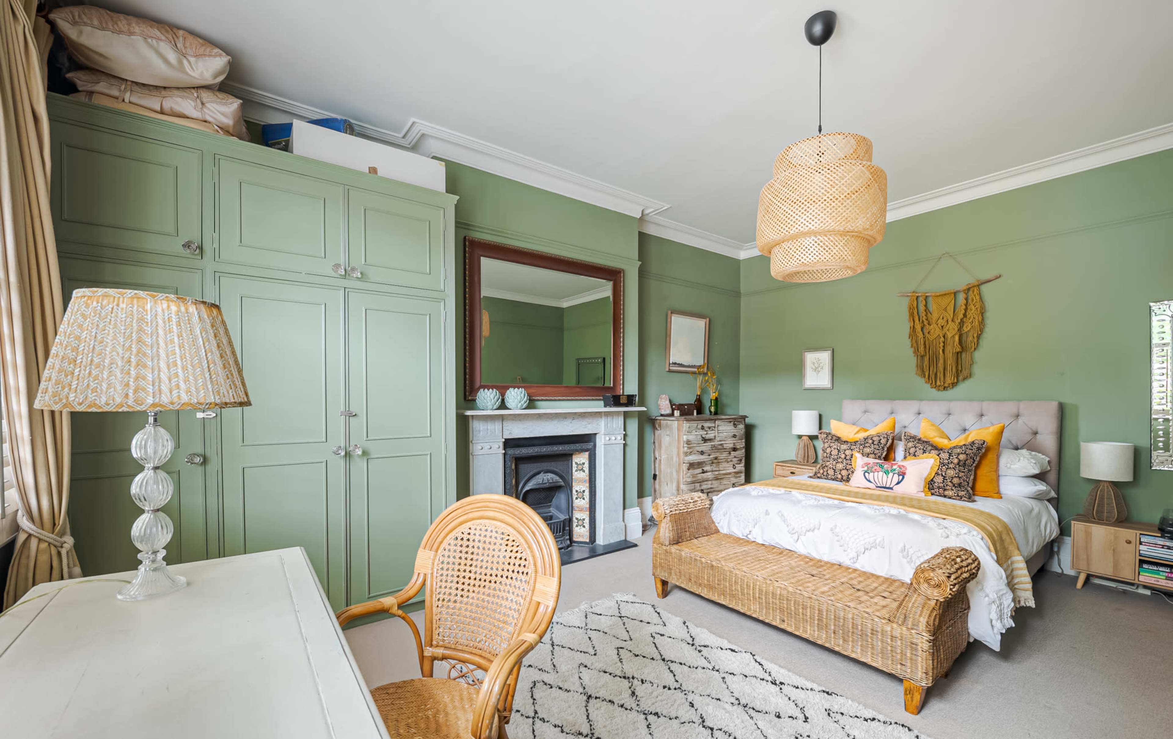 The image shows a bedroom featuring a bed with decorative pillows, a wicker bench, a desk with a chair, and a fireplace, all set against green walls.