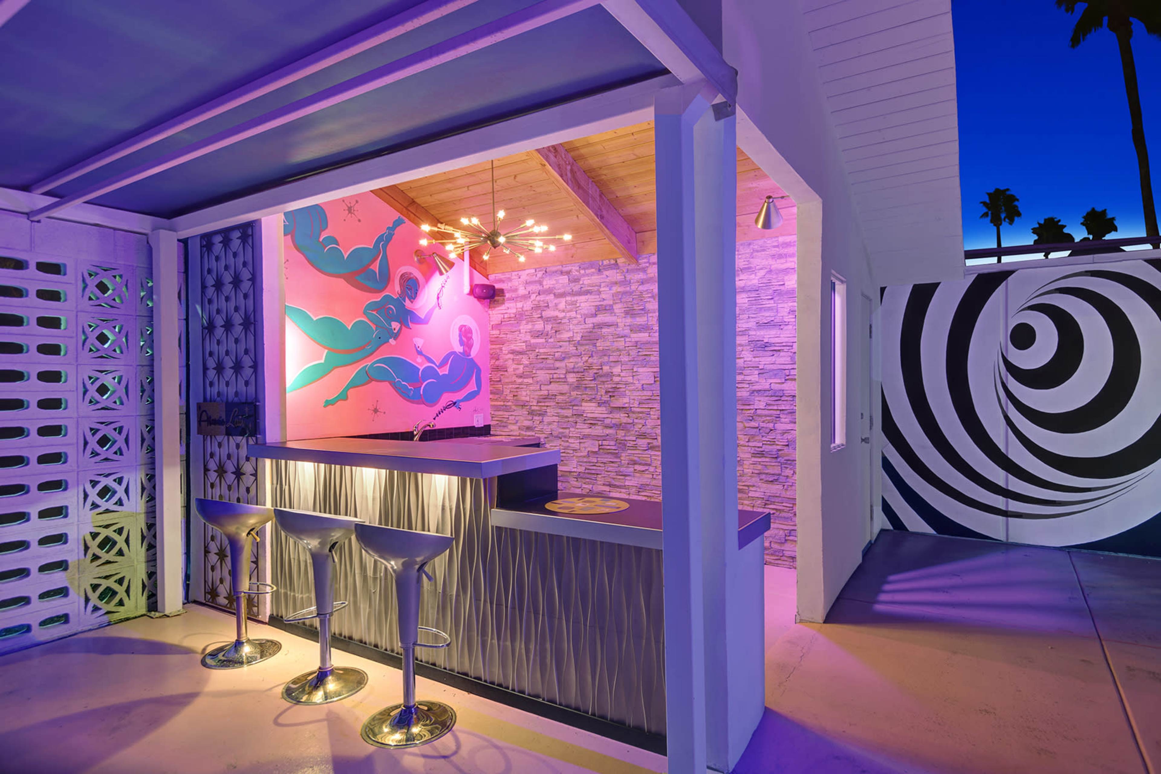 The image shows a modern outdoor bar area with two bar stools, a decorative wall mural, and colorful lighting.