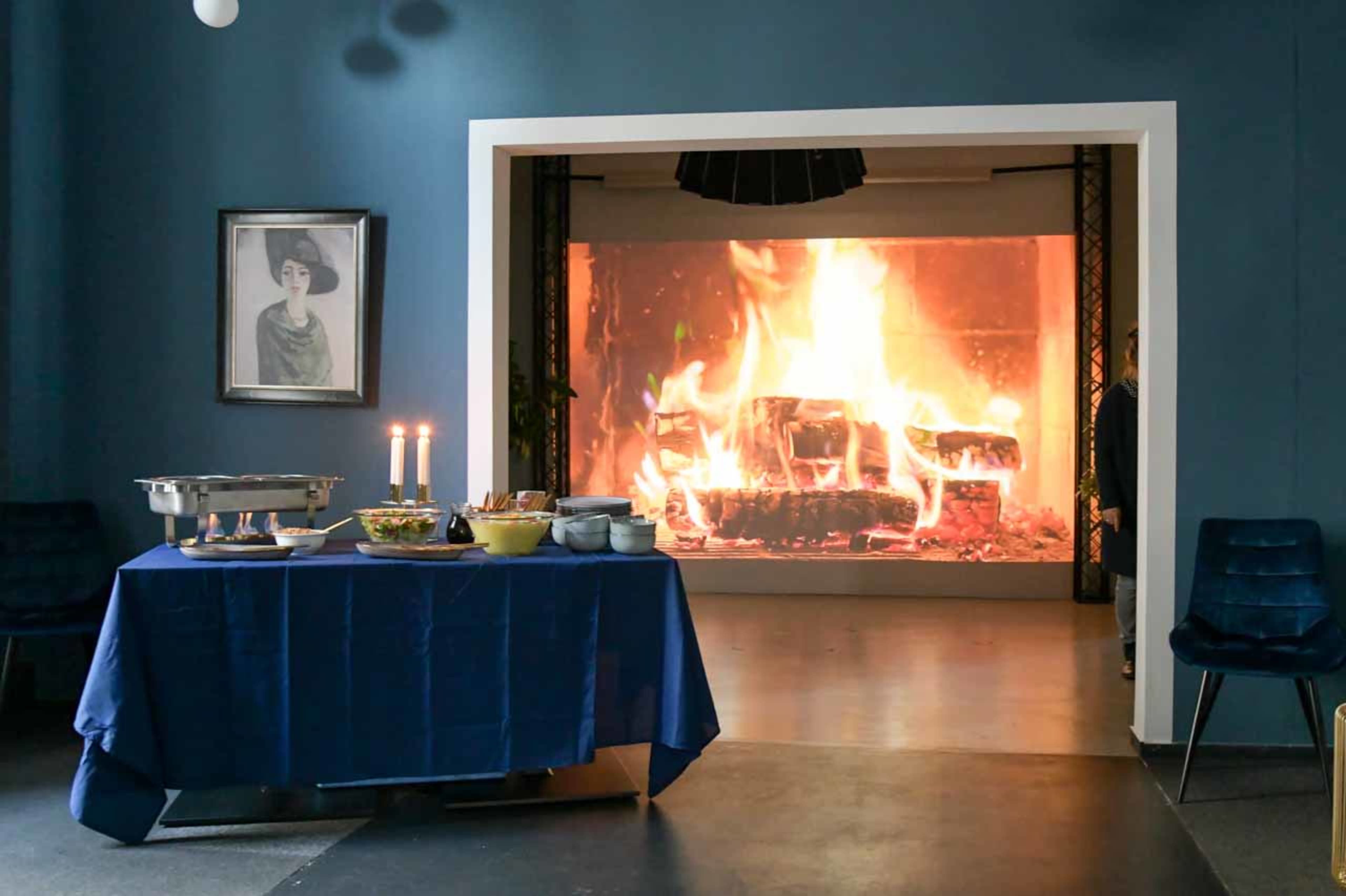 A table with food and candles is set up in front of a large screen displaying a fire.