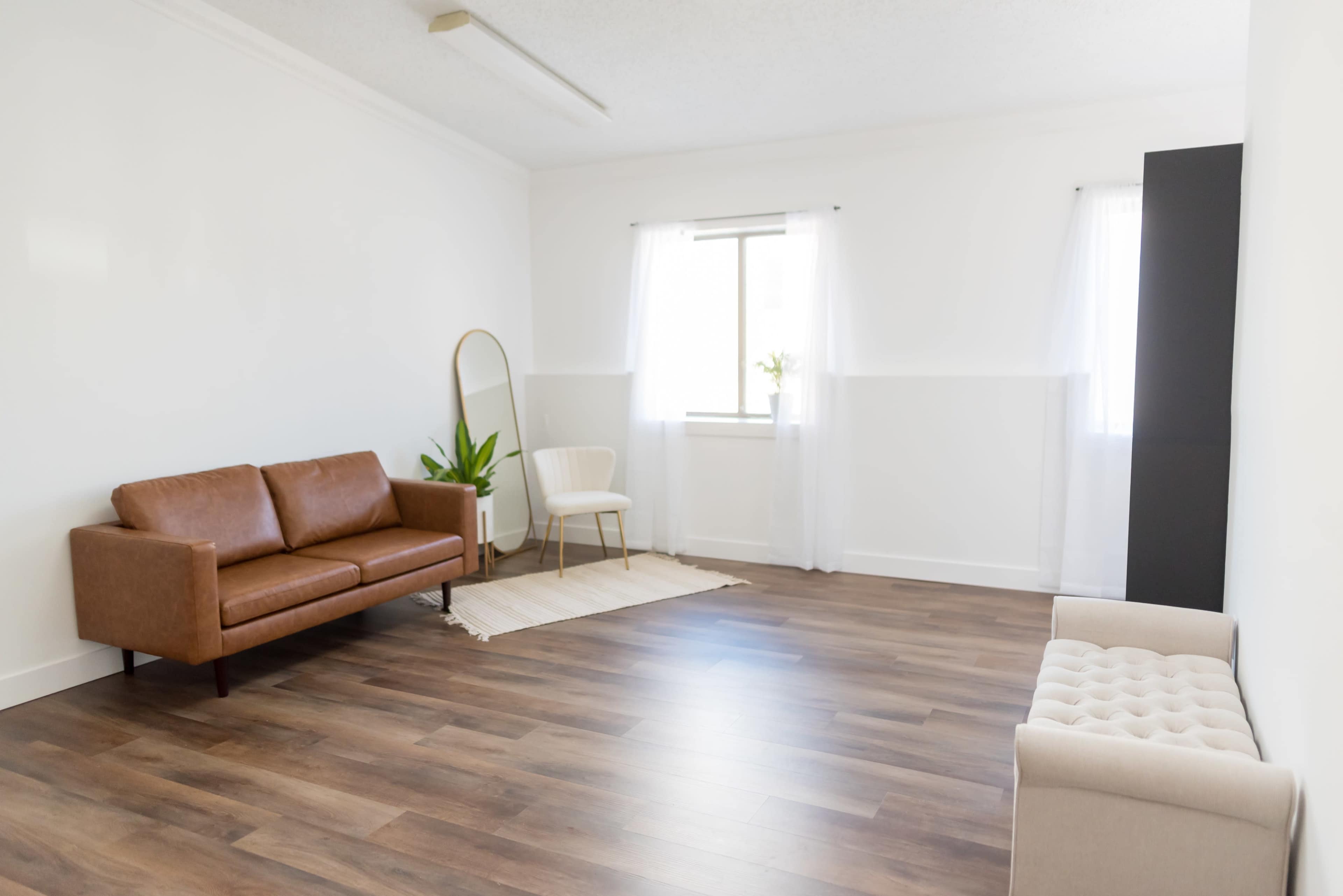 A minimalistic room with a brown leather sofa, a white chair, a large window, and a small plant, all set on a wooden floor.