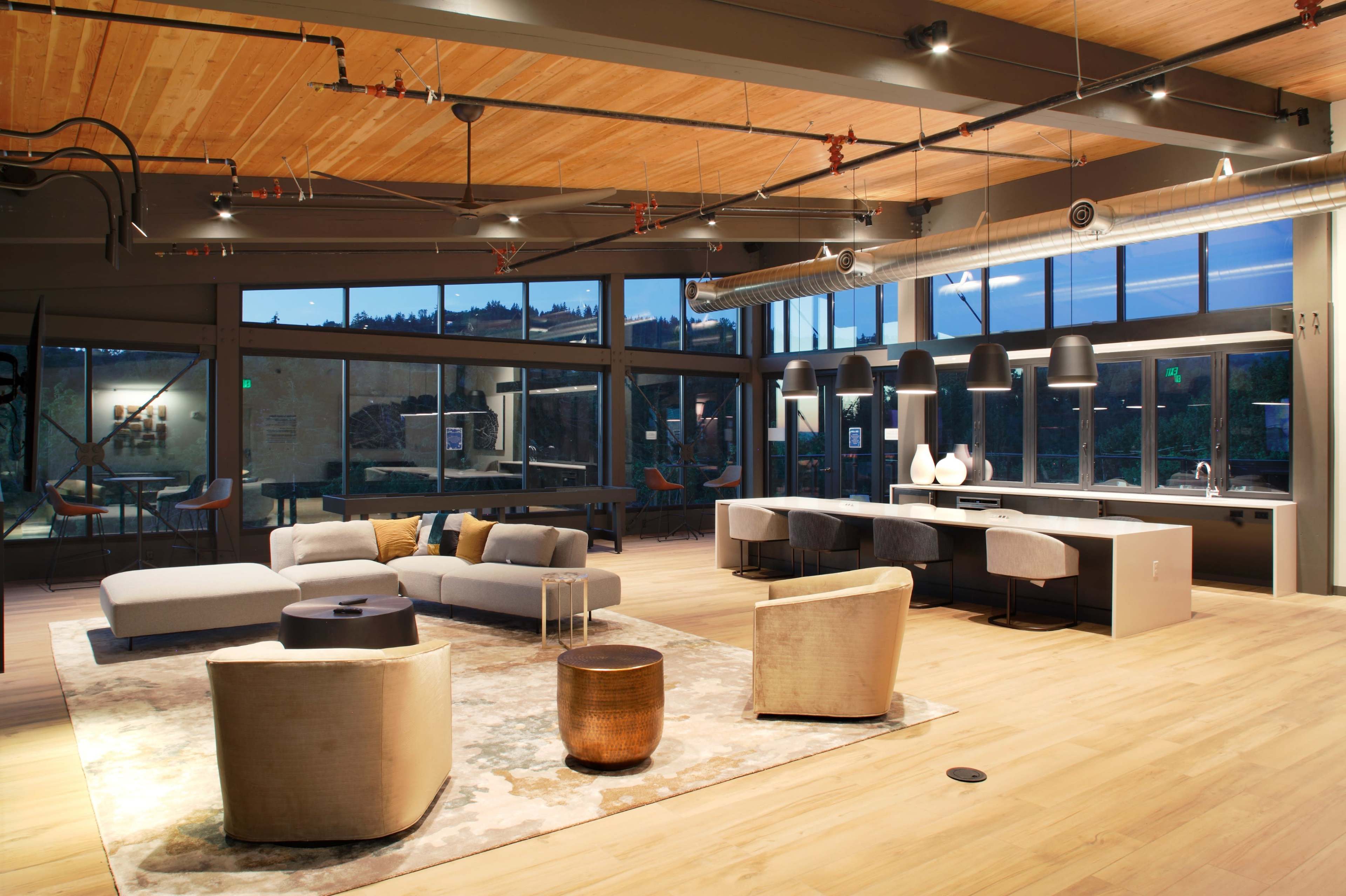 A modern, open-concept living space with a seating area featuring a sofa, armchairs, and a coffee table, alongside a kitchen area illuminated by pendant lights and large windows.
