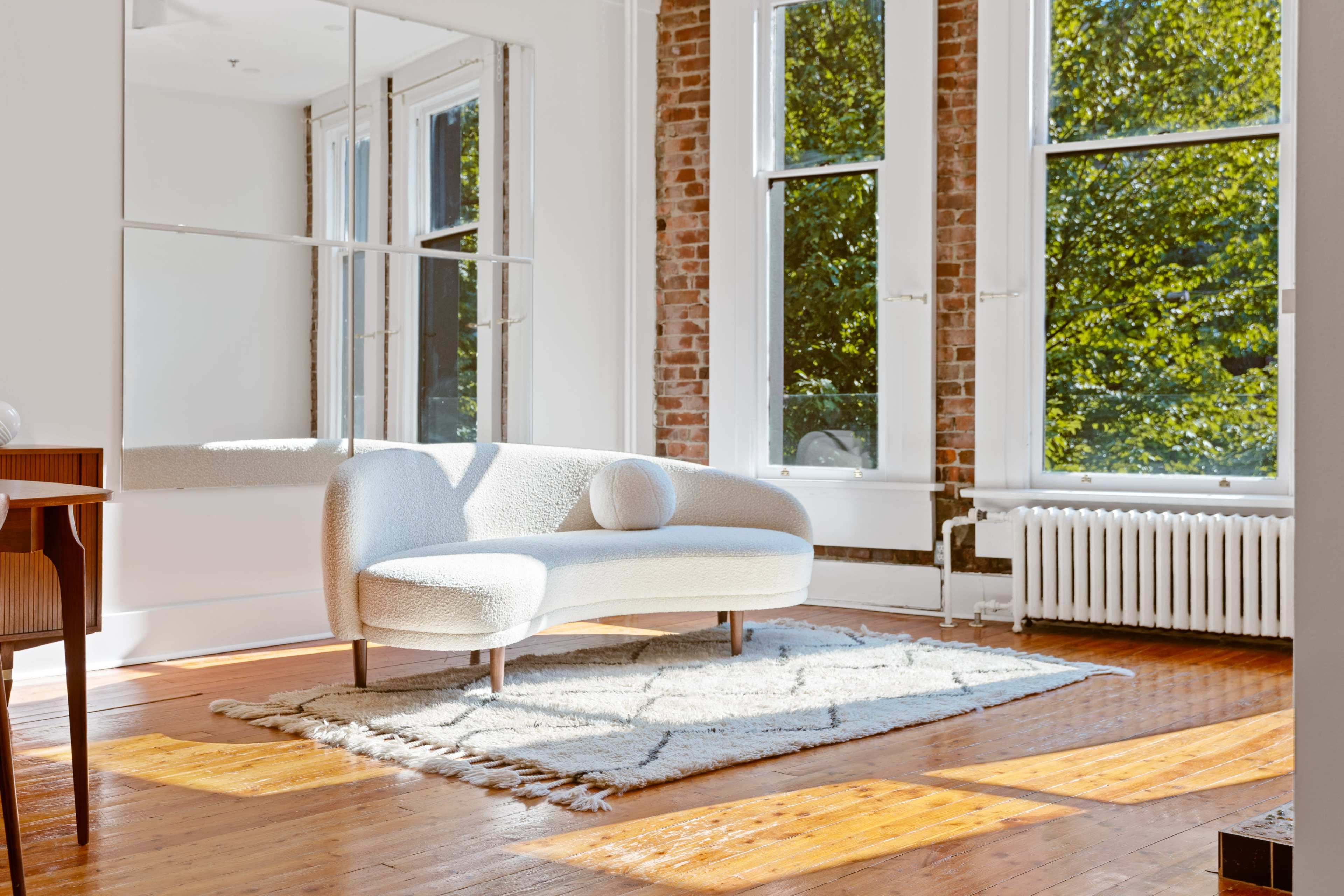 A bright living space featuring a curved white sofa on a textured rug, with large windows allowing natural light to illuminate the room and expose brick walls.