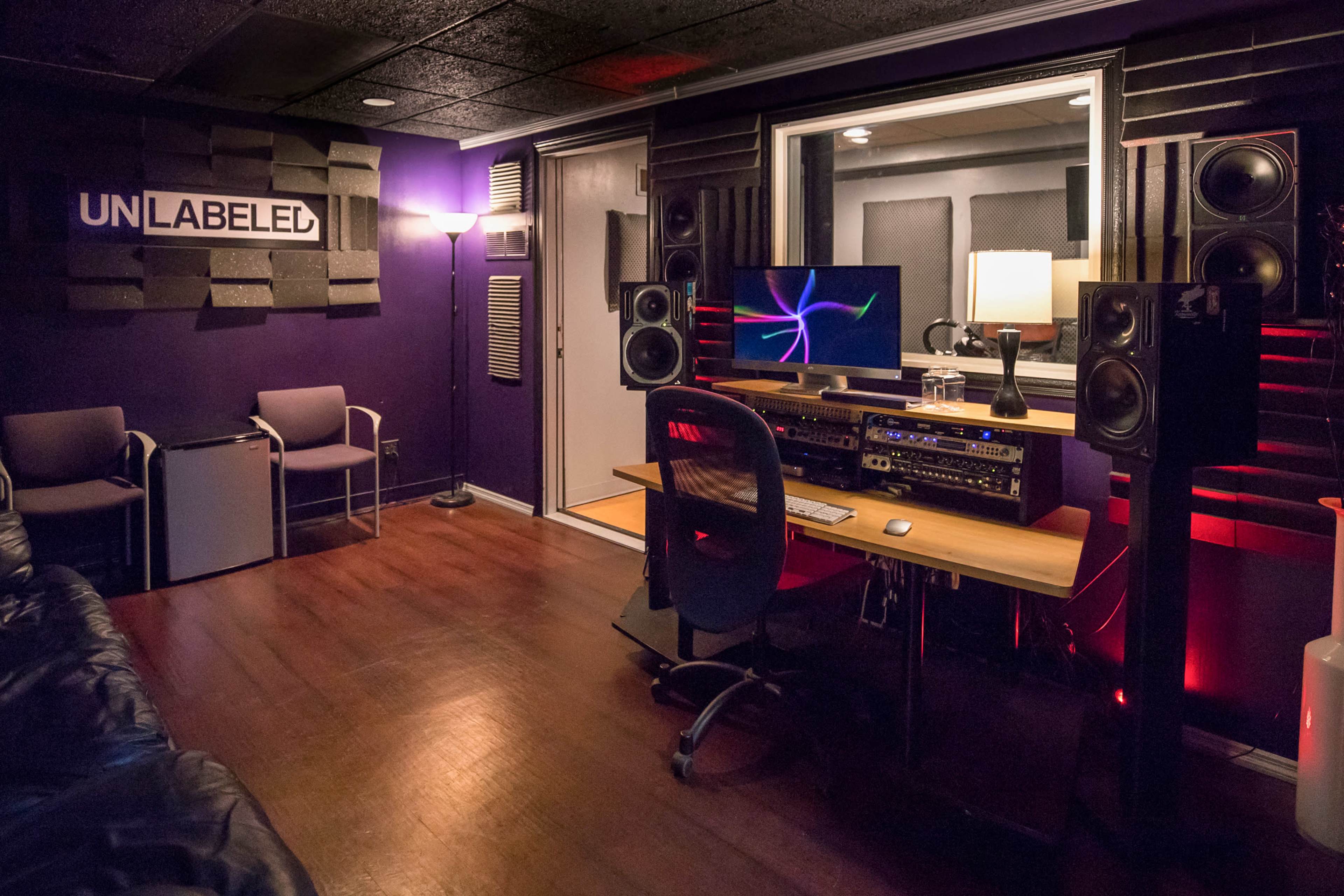 A modern music studio featuring a control desk with audio equipment, a computer monitor, two large speakers, and a seating area with chairs against a dark purple backdrop.
