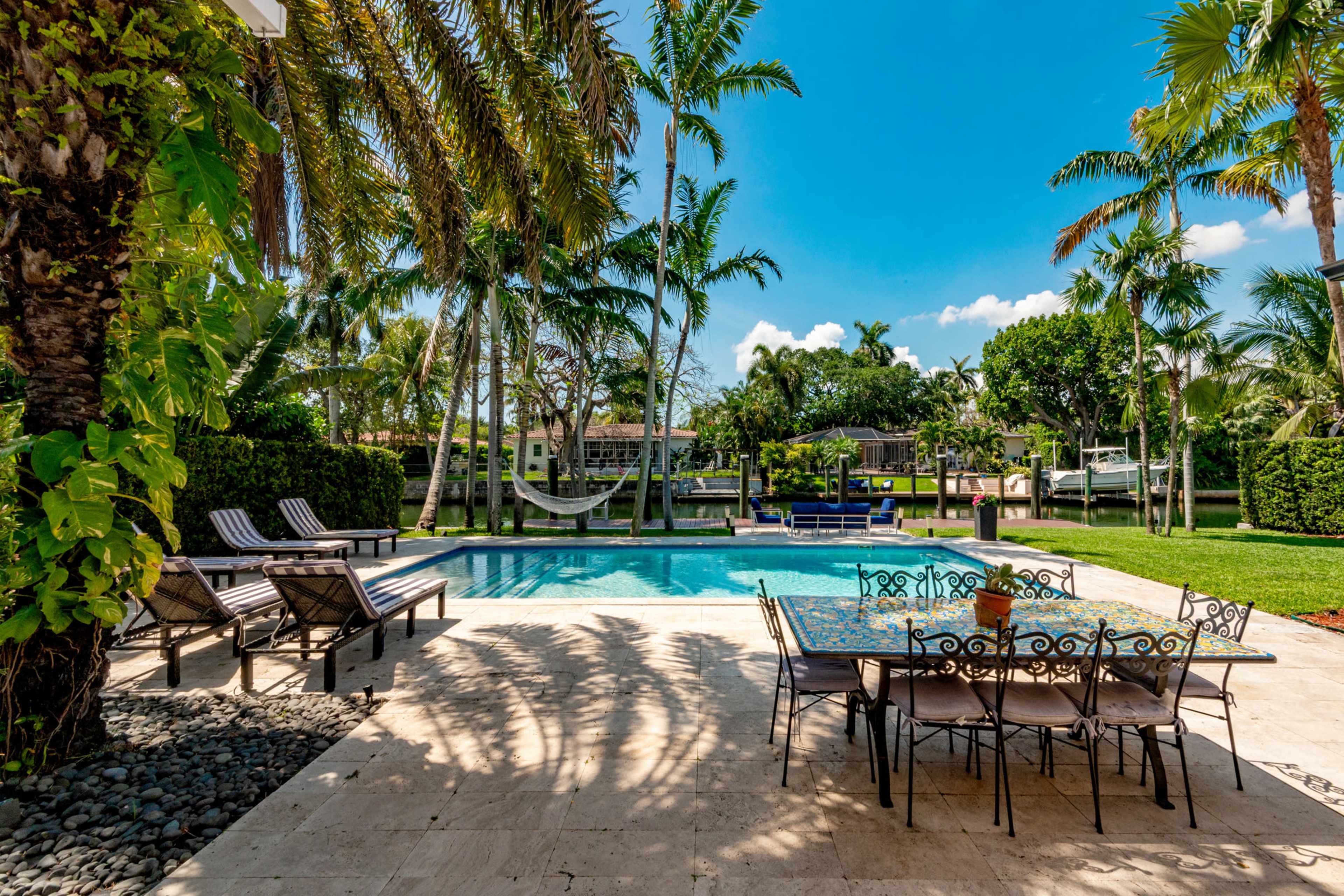 A pool area with lounge chairs and a dining set is surrounded by palm trees and greenery under a clear blue sky.