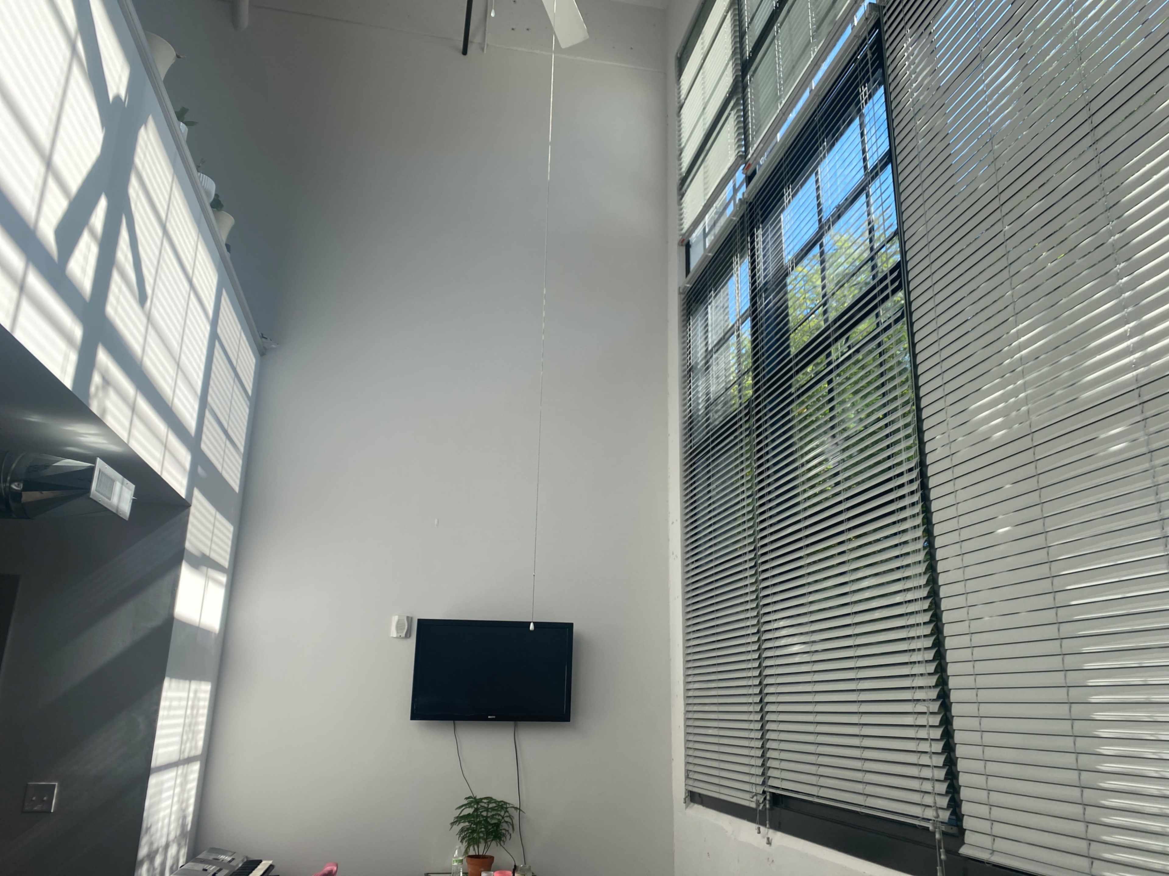 A room with a high ceiling, a television mounted on a light-colored wall, and large windows featuring blinds.