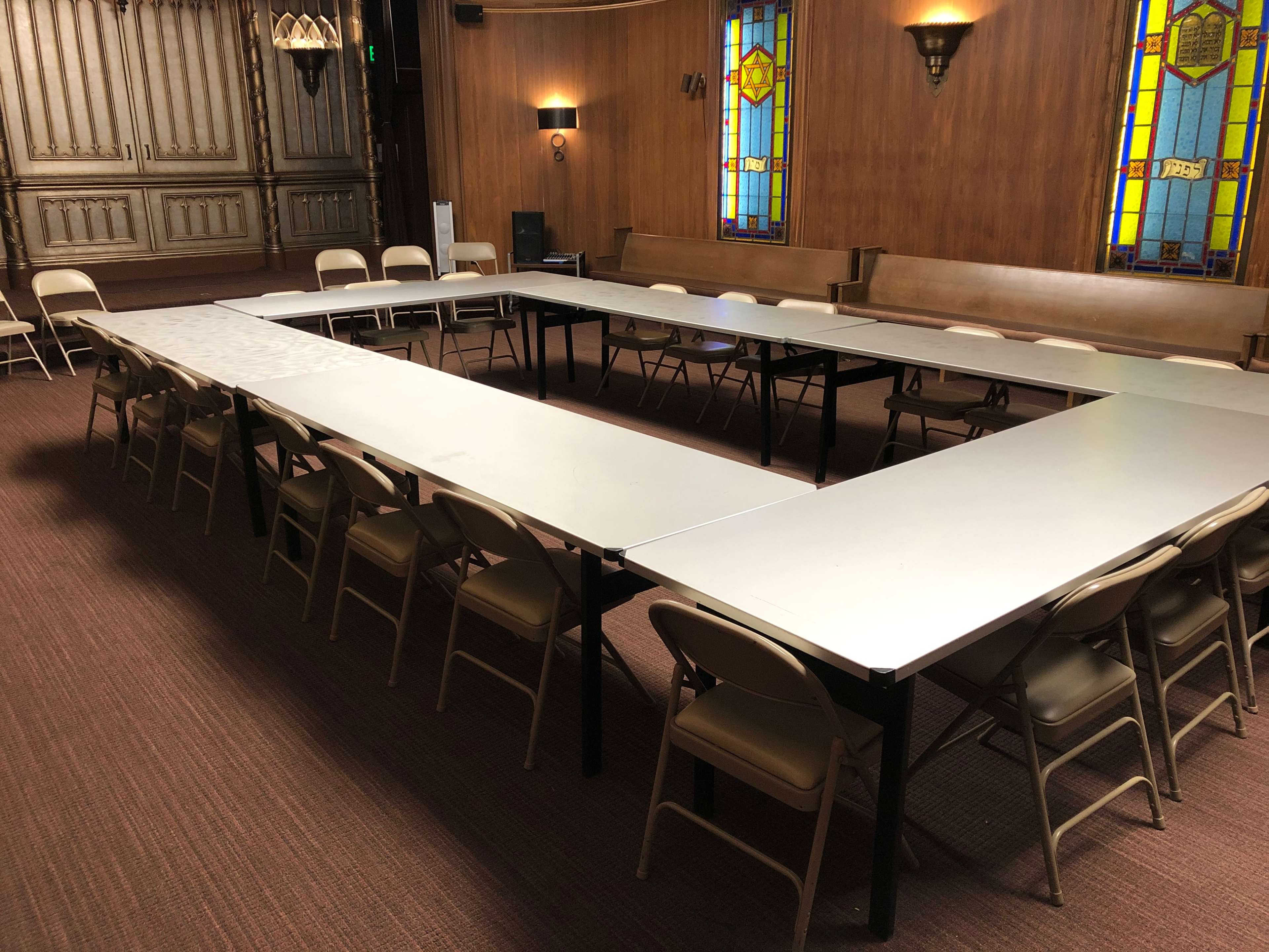 A large meeting room features an arrangement of tables in a U-shape, surrounded by folding chairs, with stained glass windows in the background.