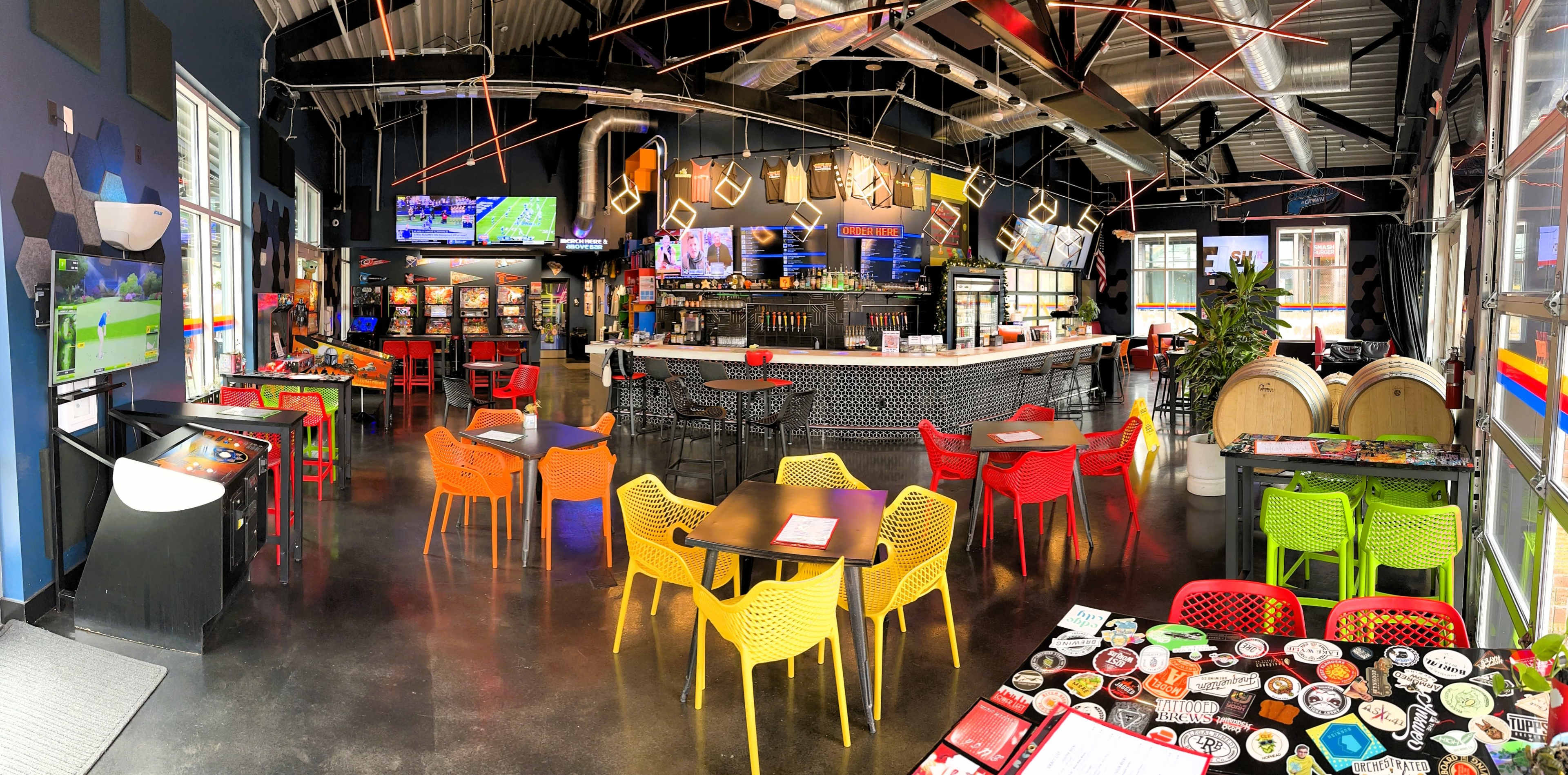 A modern bar and arcade with colorful chairs, a long counter, and various arcade machines and TVs displaying sports.