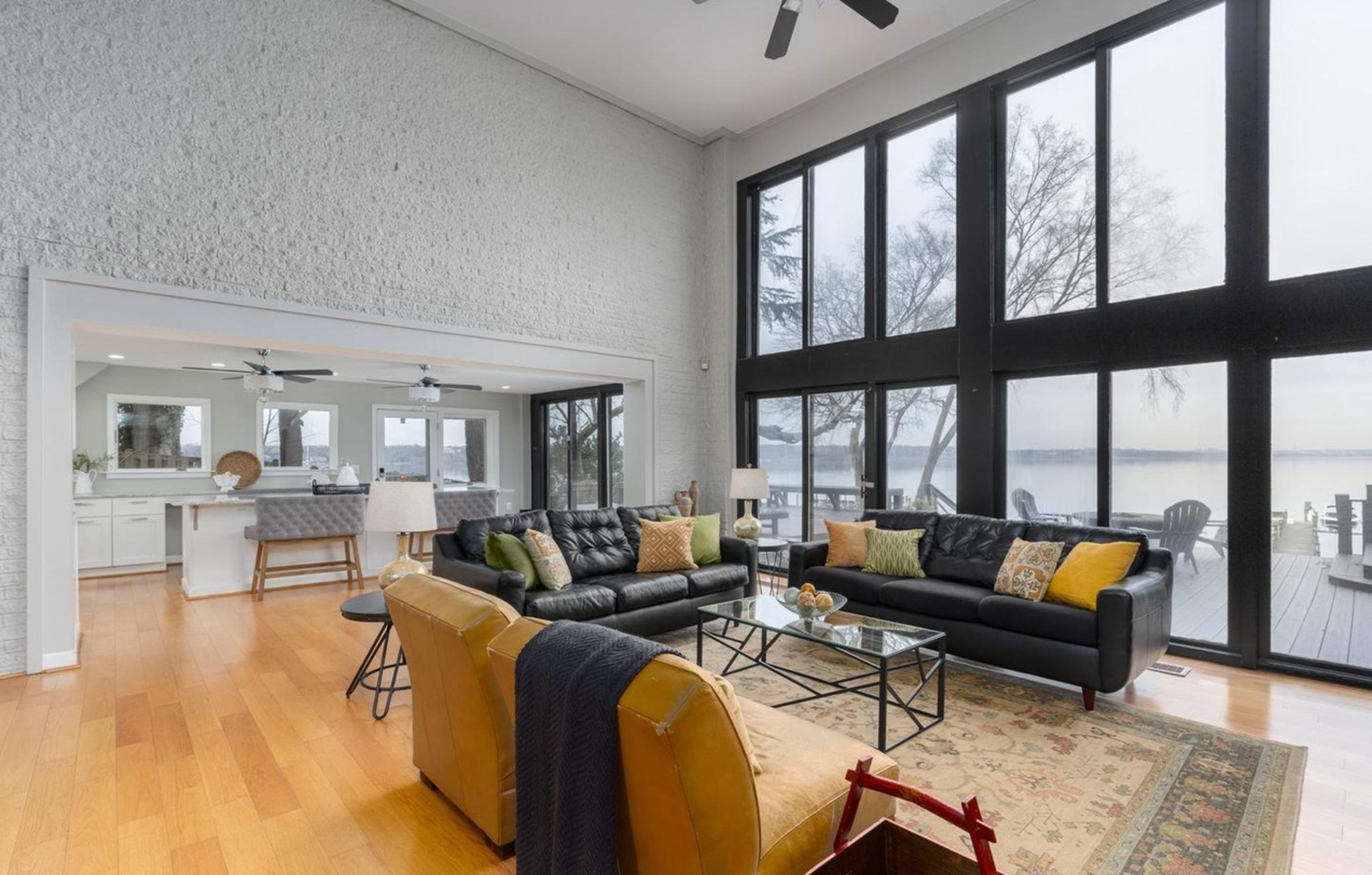 The spacious living room features large windows that provide a view of the lake, complemented by black leather sofas, a glass coffee table, and a mix of decorative cushions.