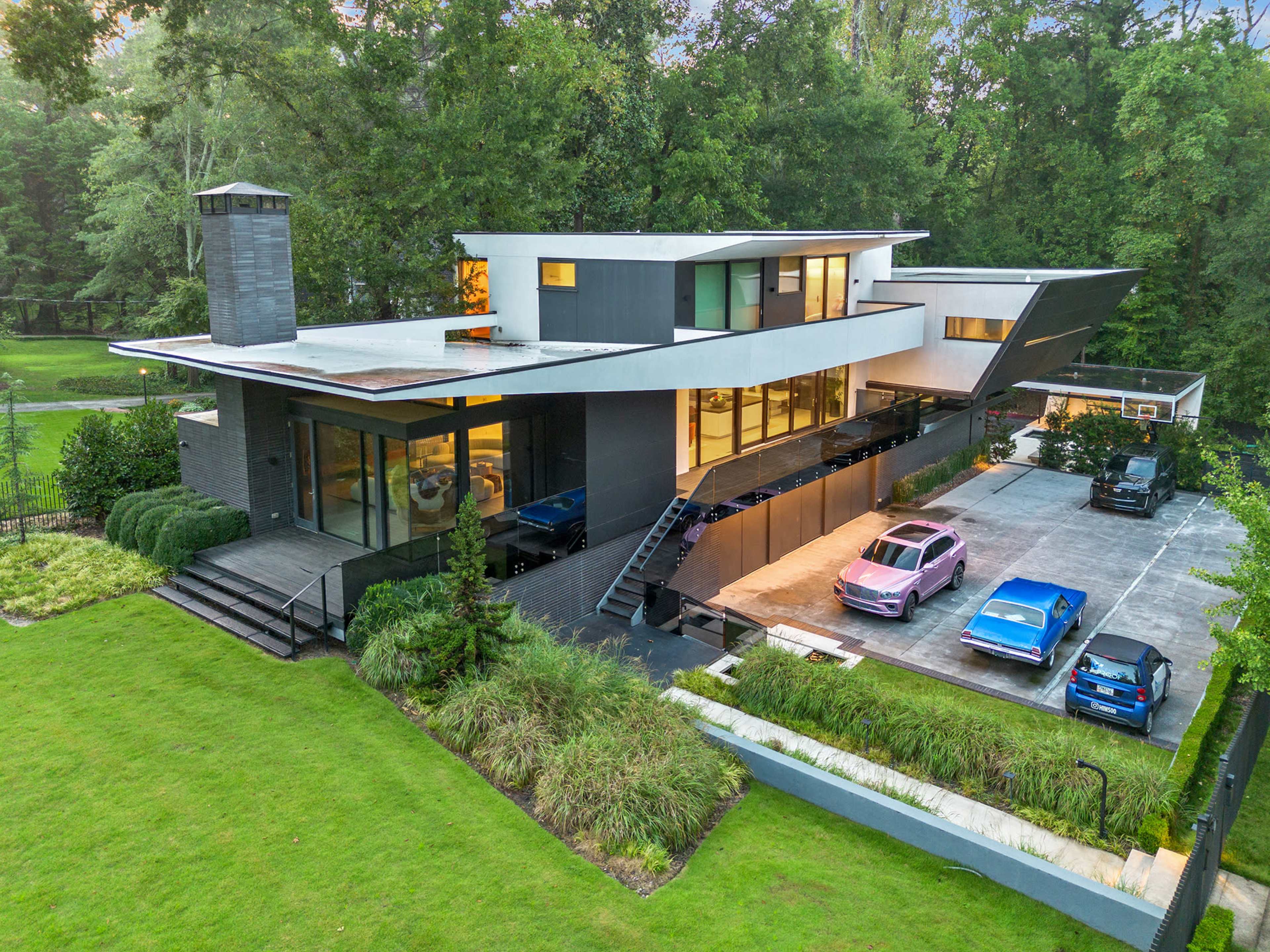 A modern two-story house features a unique angular design, large windows, and a driveway with multiple parked cars, set against a lush green landscape.