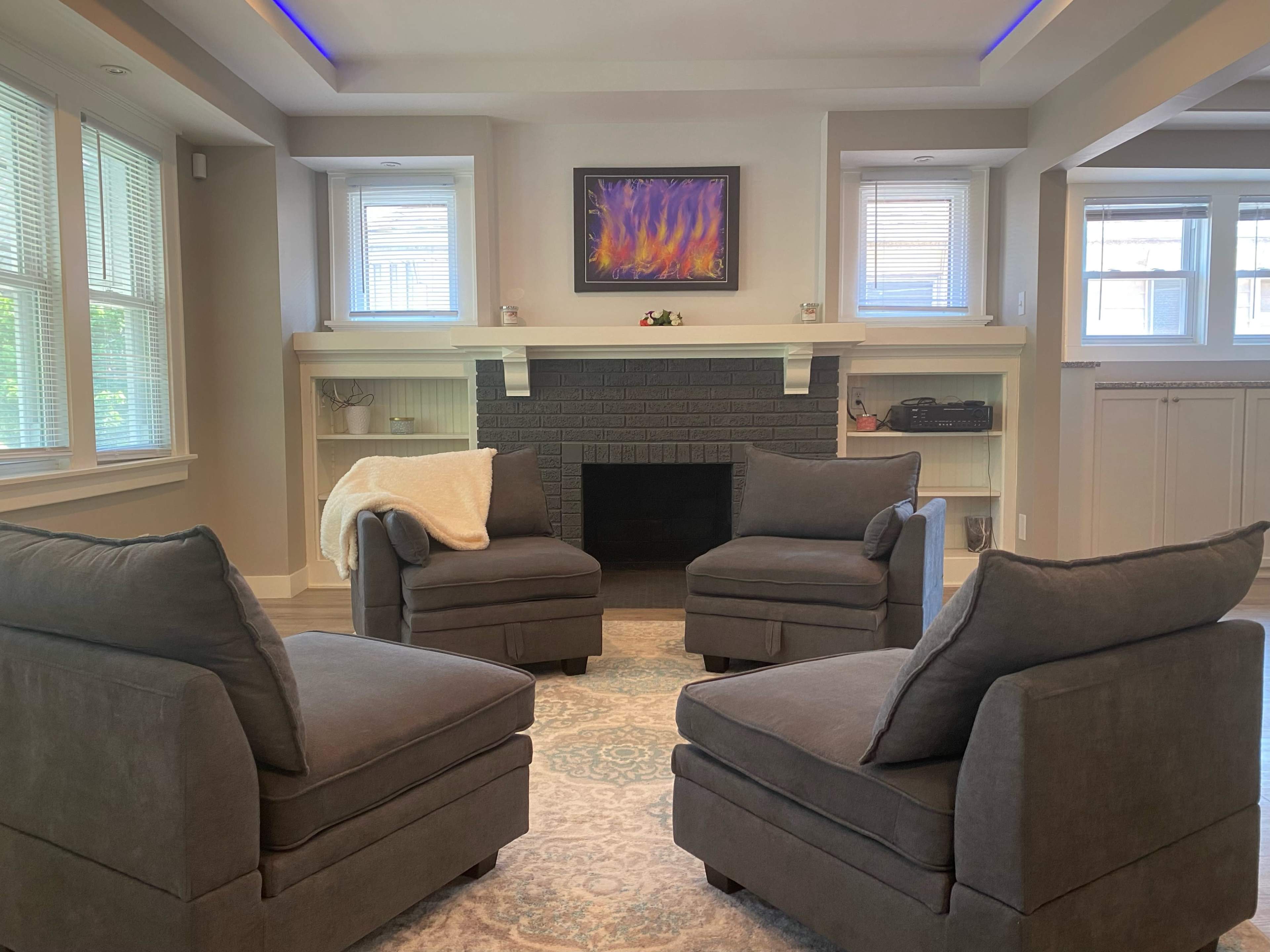 A living room with four cushioned chairs arranged around a low table near a dark fireplace, with windows allowing natural light and a colorful painting above the mantel.