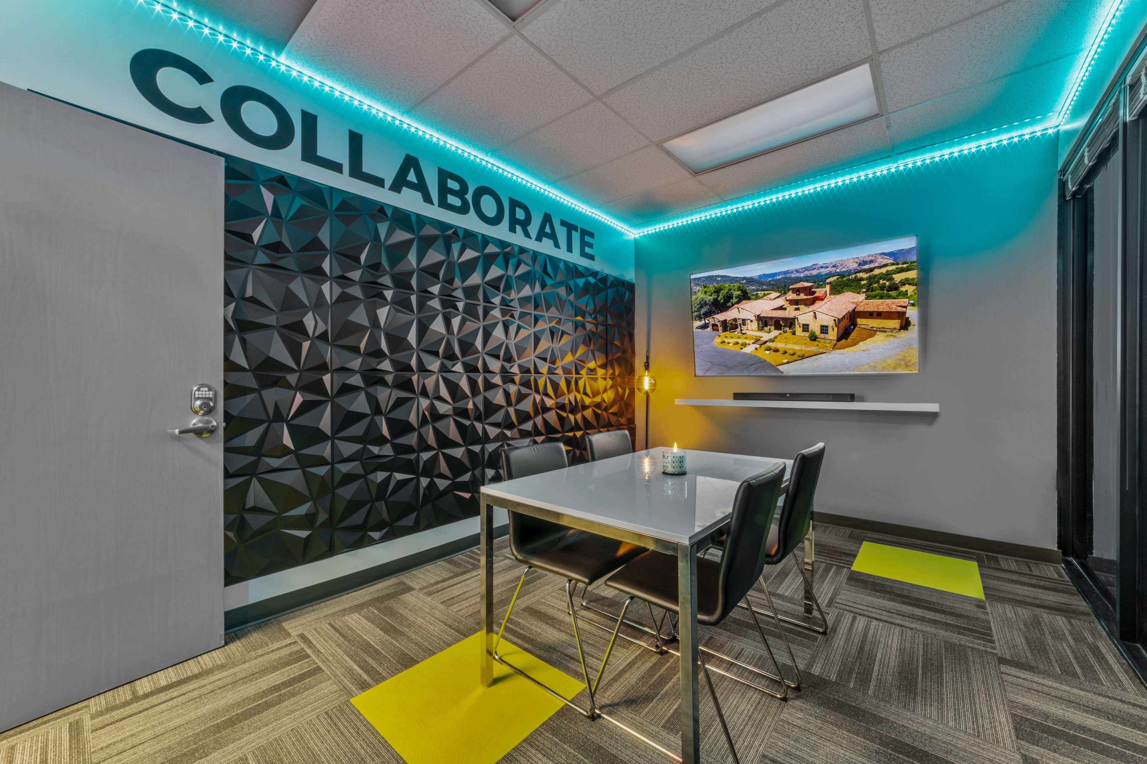 A modern meeting room features a table with chairs, a large wall-mounted screen, and a textured black accent wall with the word "COLLABORATE" displayed.