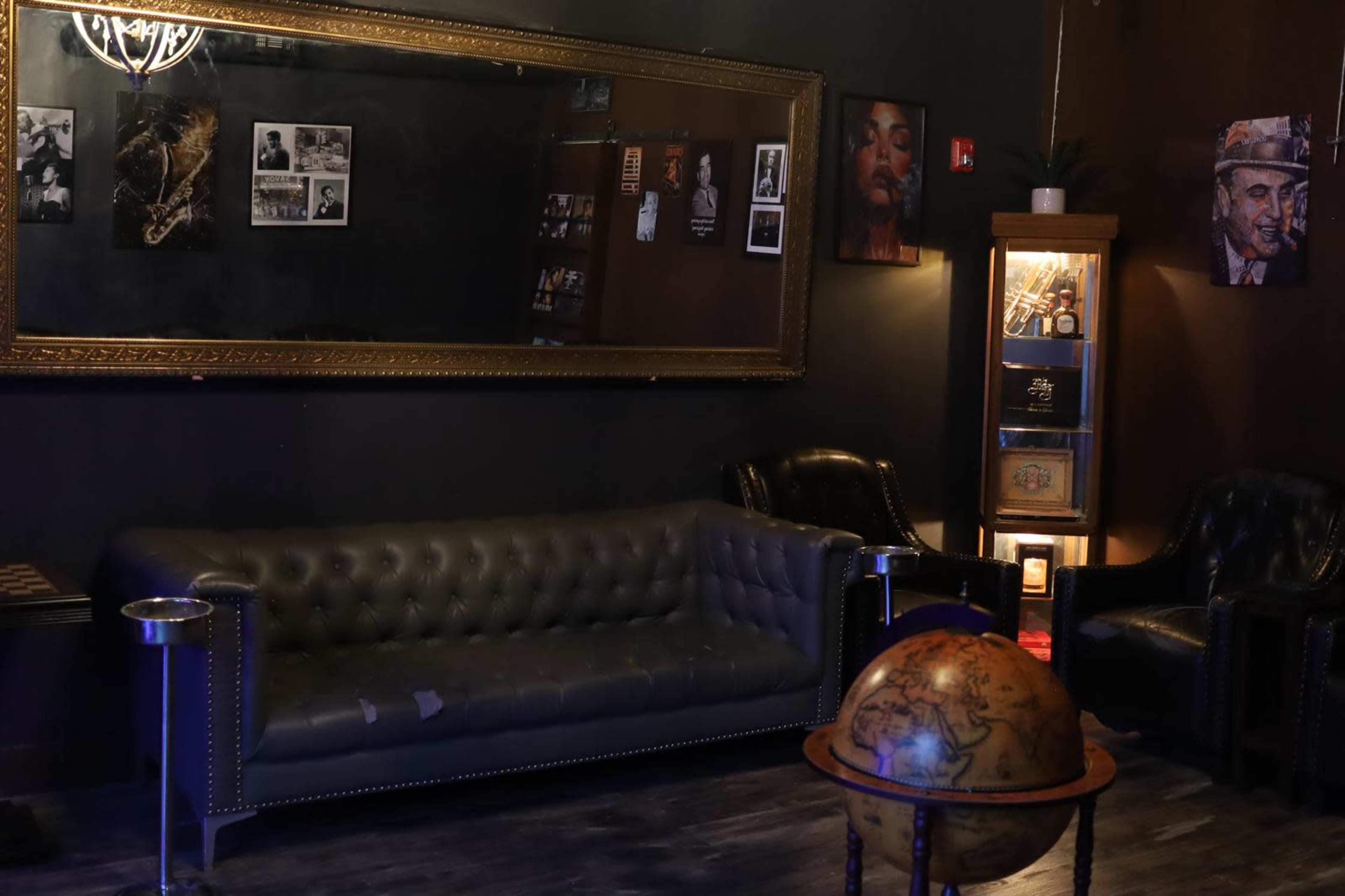 The image shows a dimly lit lounge area featuring a leather sofa, two armchairs, a globe, a large mirror, and framed photographs on the walls.