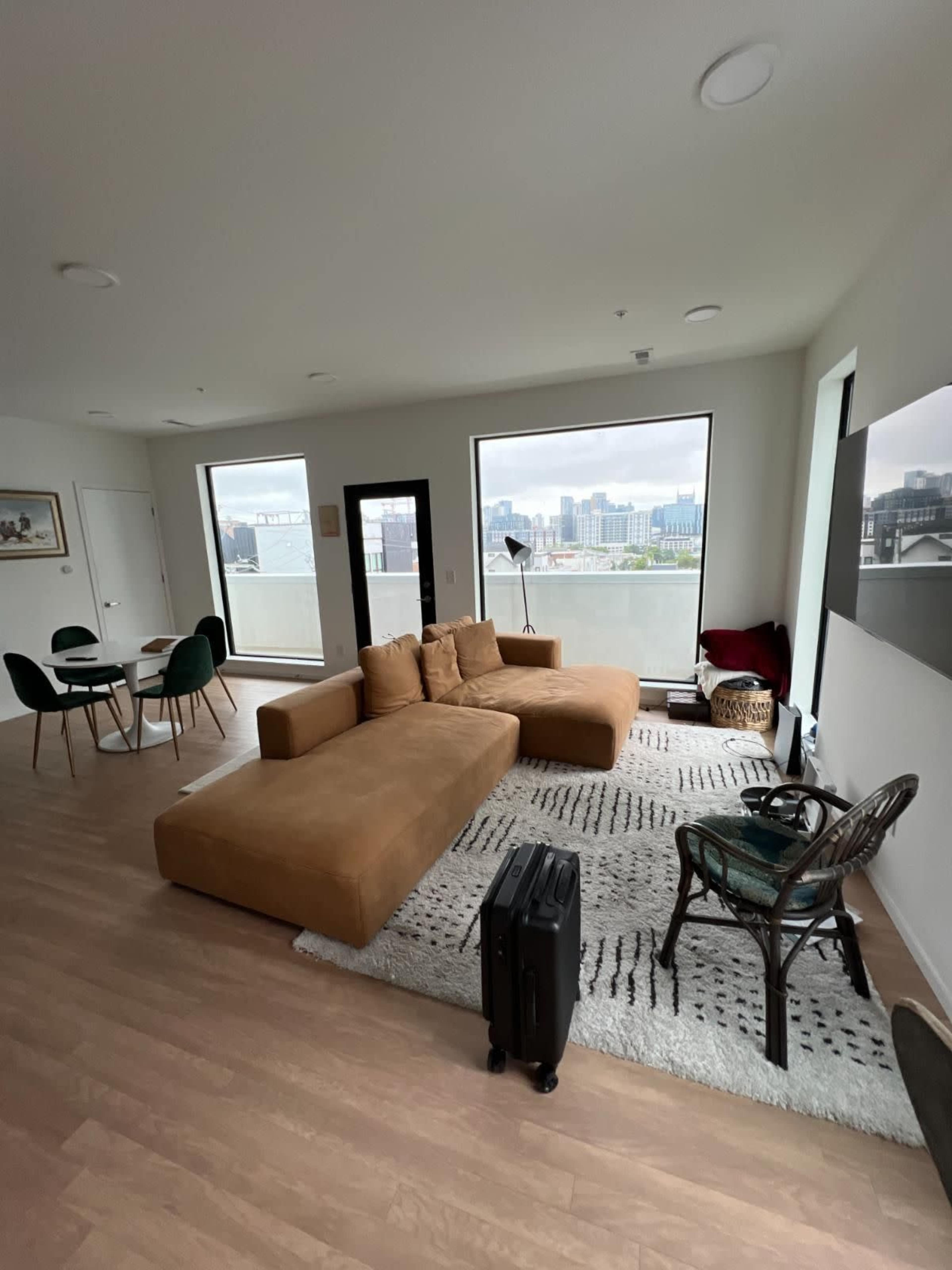 A modern living area featuring a large brown sectional sofa, a circular dining table with green chairs, a suitcase, and large windows showcasing a city skyline.