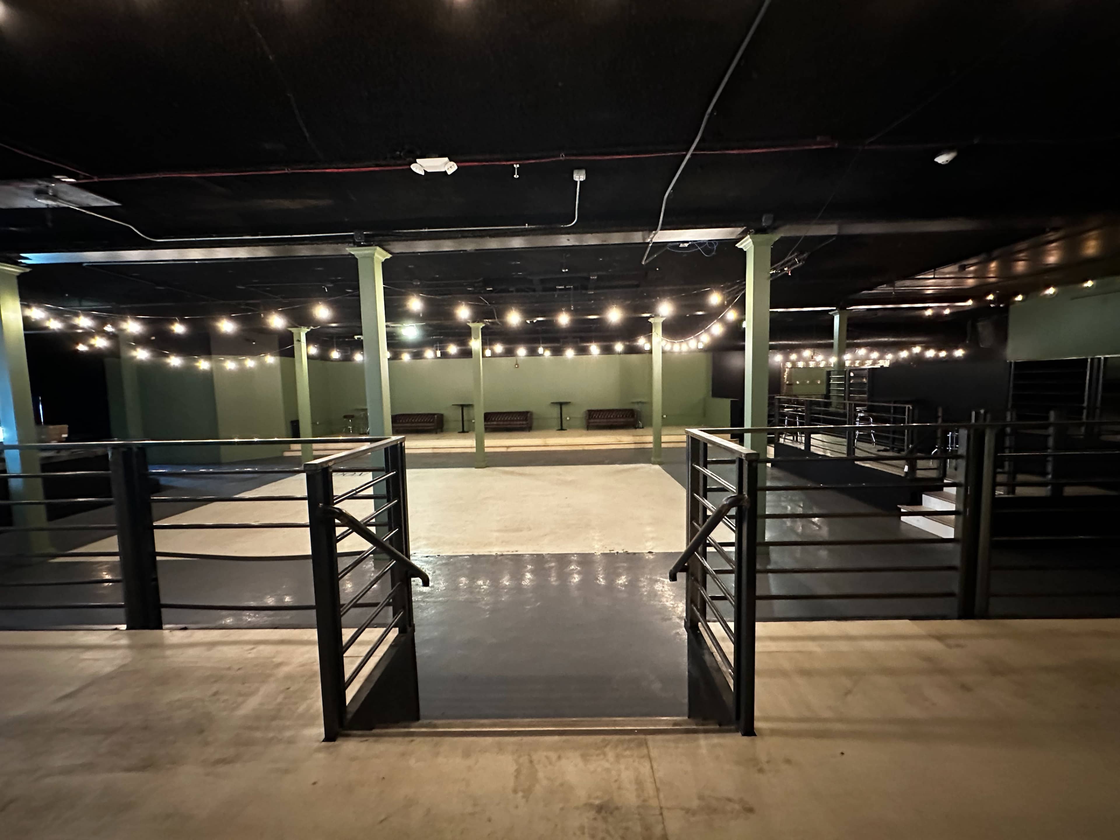 A dimly lit event space with string lights overhead, featuring a large open floor and railing on either side.