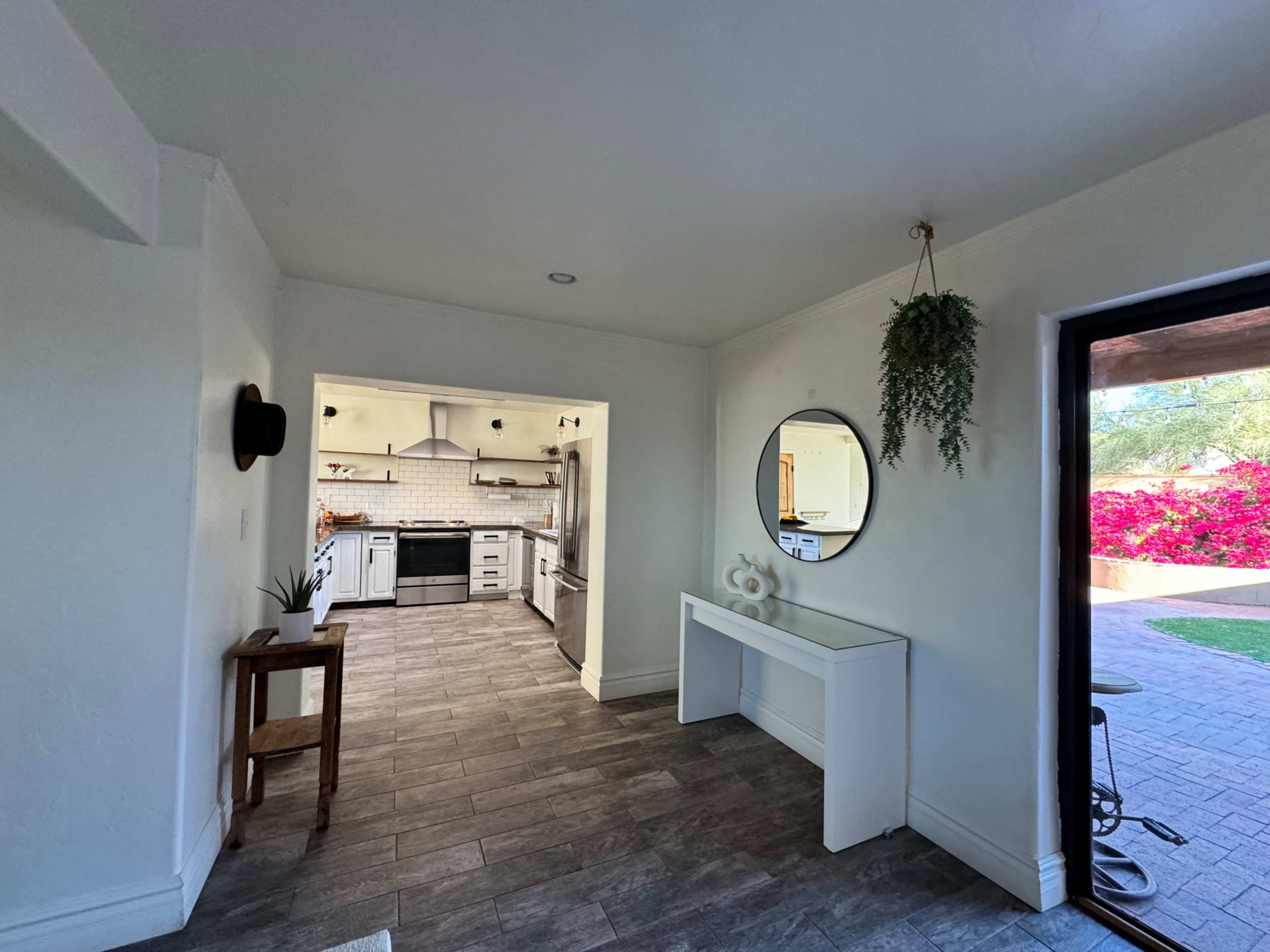 The image shows a bright, open foyer leading to a kitchen, featuring a round mirror and a small table on one side.