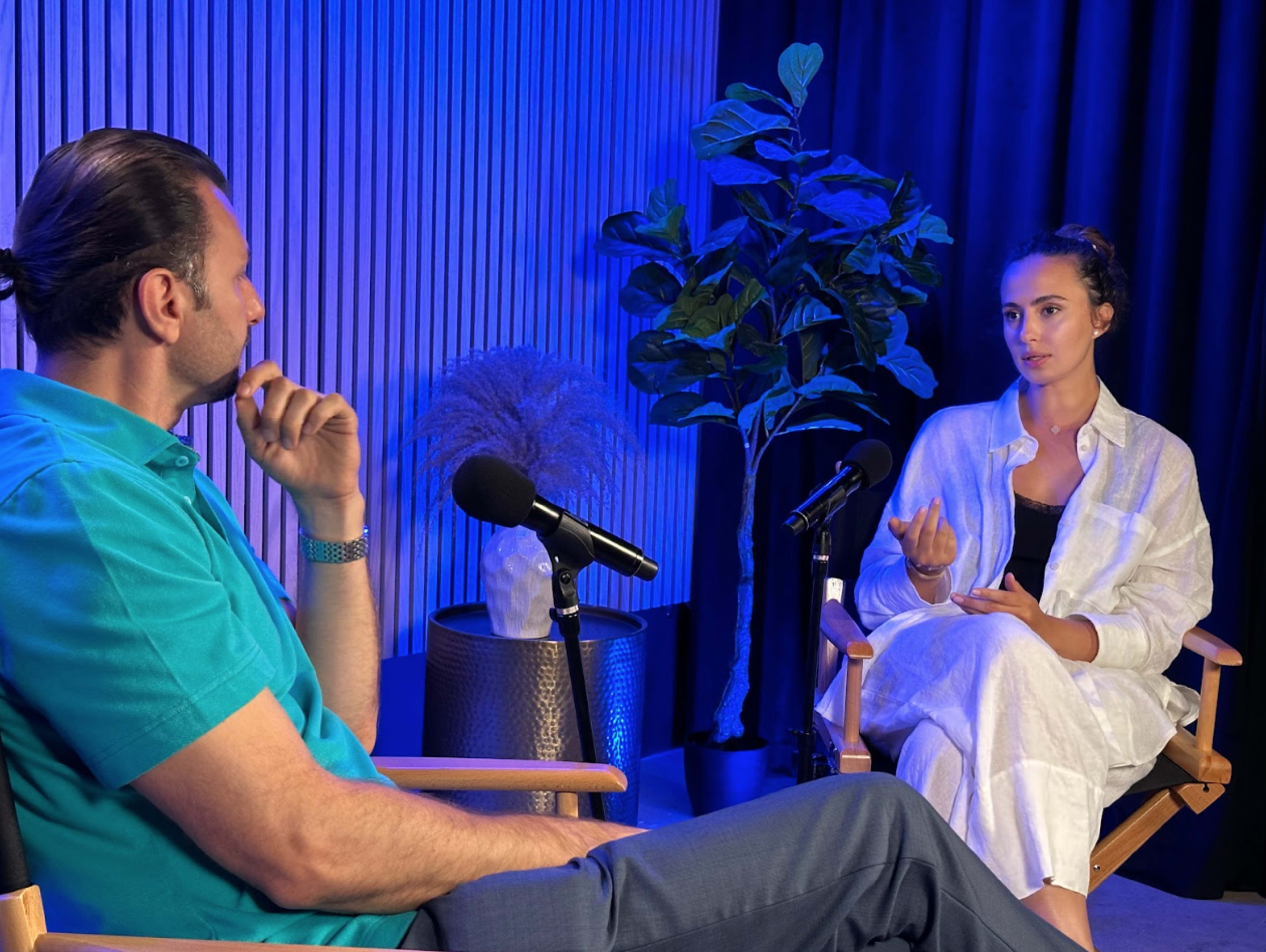 A man and a woman are seated in a studio setting, engaged in conversation, with microphones in front of them and plants in the background.