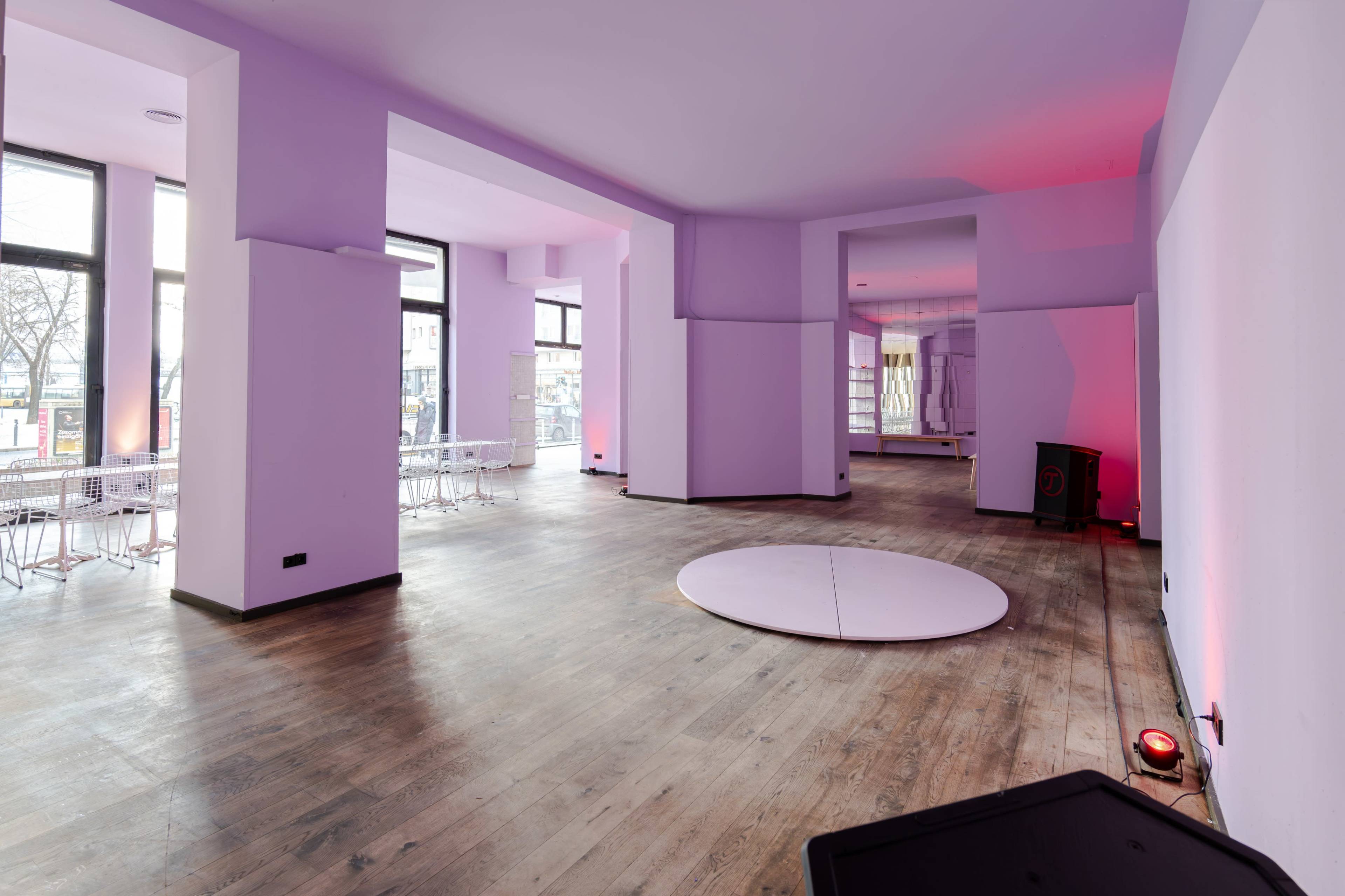 The interior features a spacious, empty room with light purple walls, wooden flooring, and a circular platform at the center.