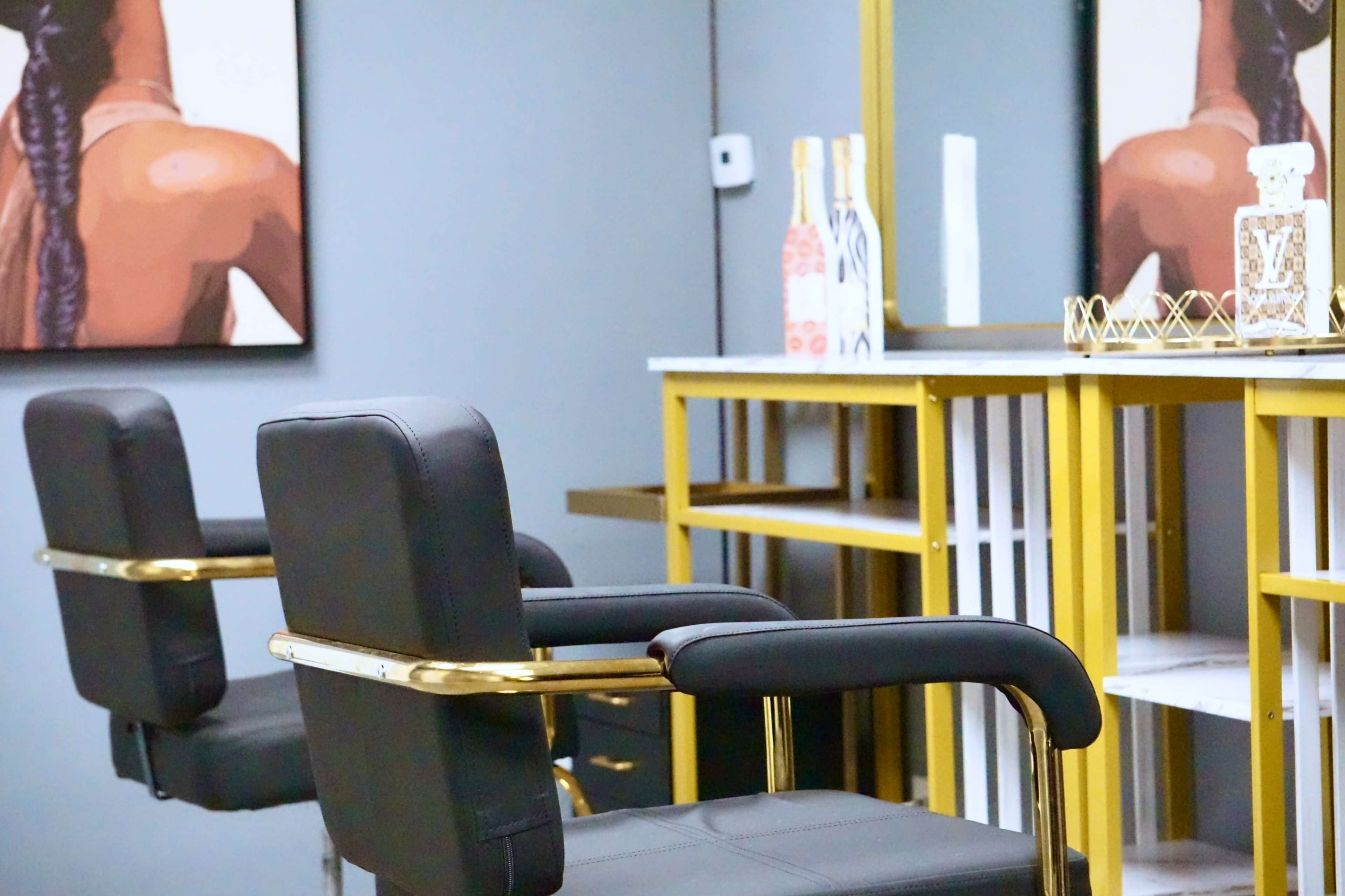 The image shows a modern salon interior with two black chairs positioned at a stylish gold-accented workstation beneath wall art depicting a woman's back.