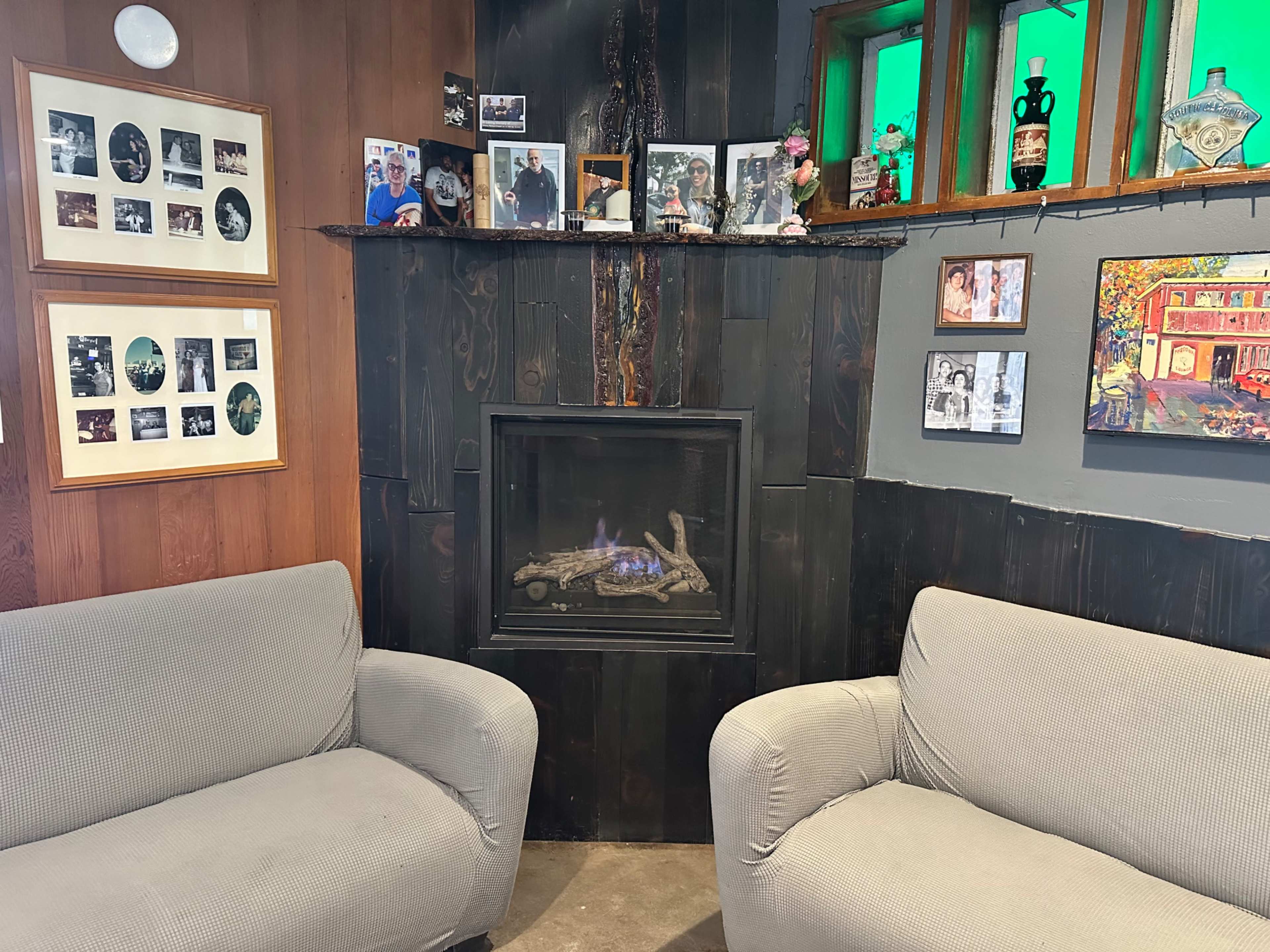The image shows a cozy corner with two grey armchairs facing a fireplace surrounded by framed photographs and artwork on the walls.