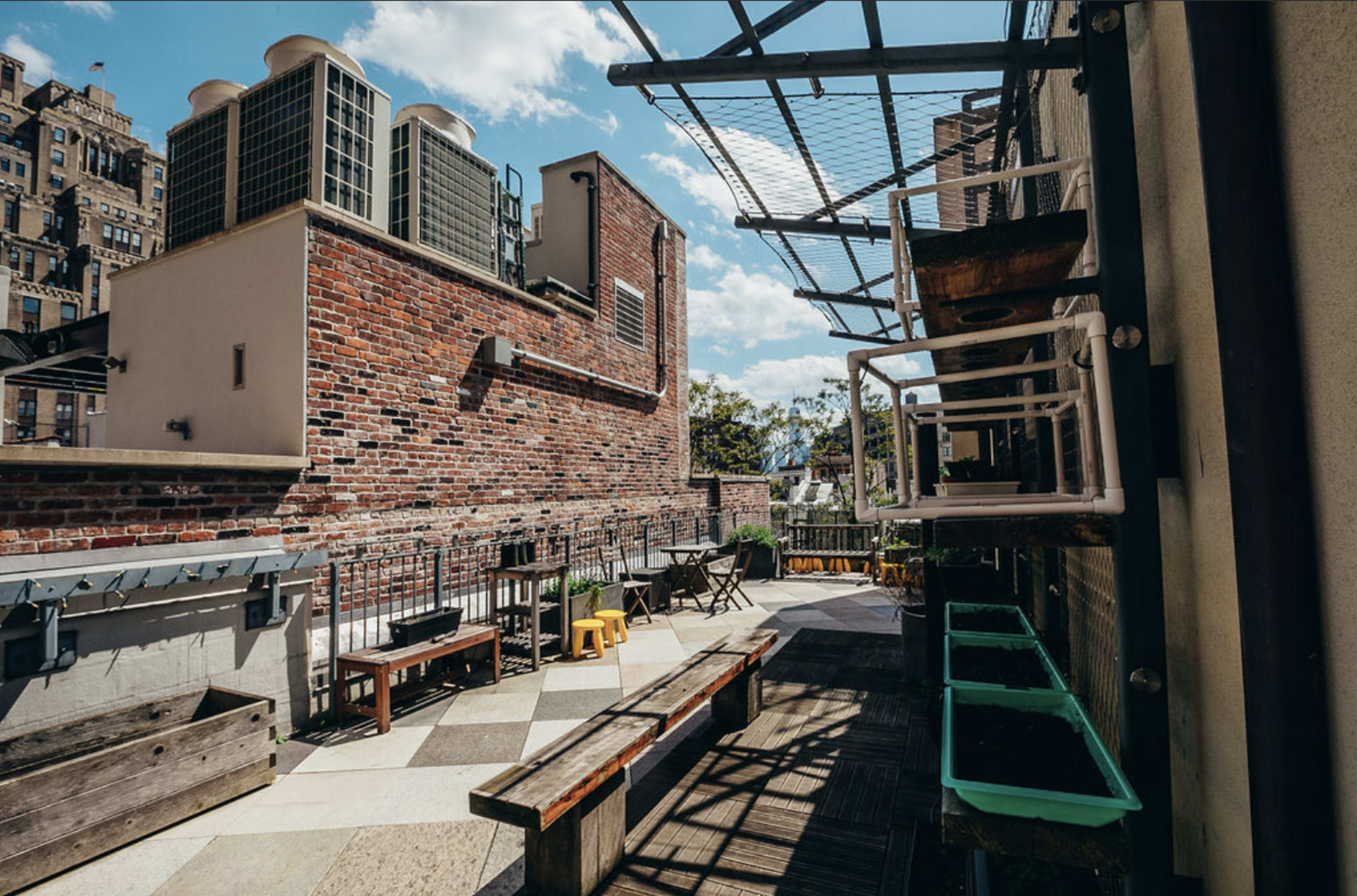 A rooftop terrace features wooden seating, gardening boxes, and a view of nearby buildings under a clear blue sky.