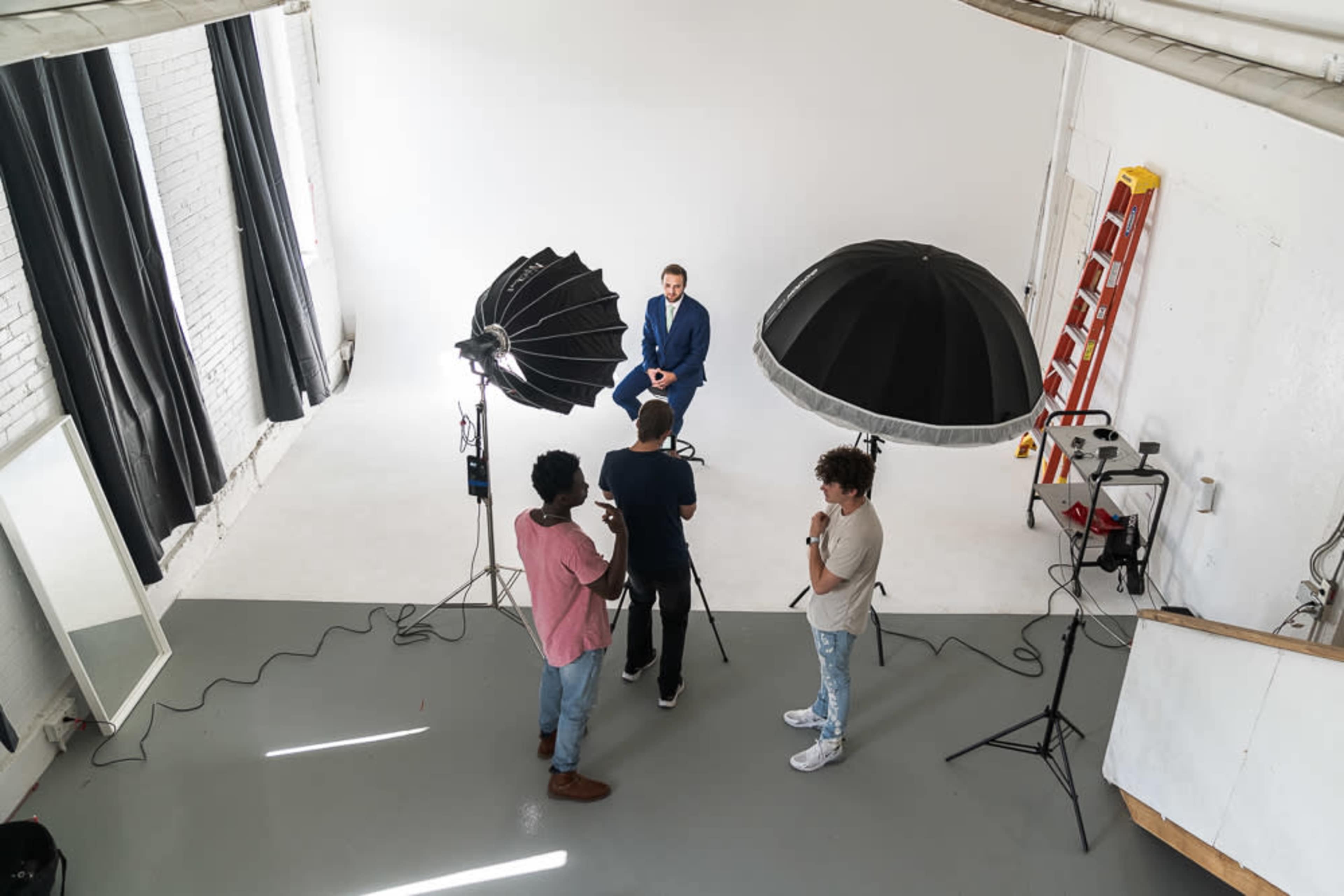A man in a blue suit is seated for a photoshoot while two other individuals set up equipment in a photography studio.