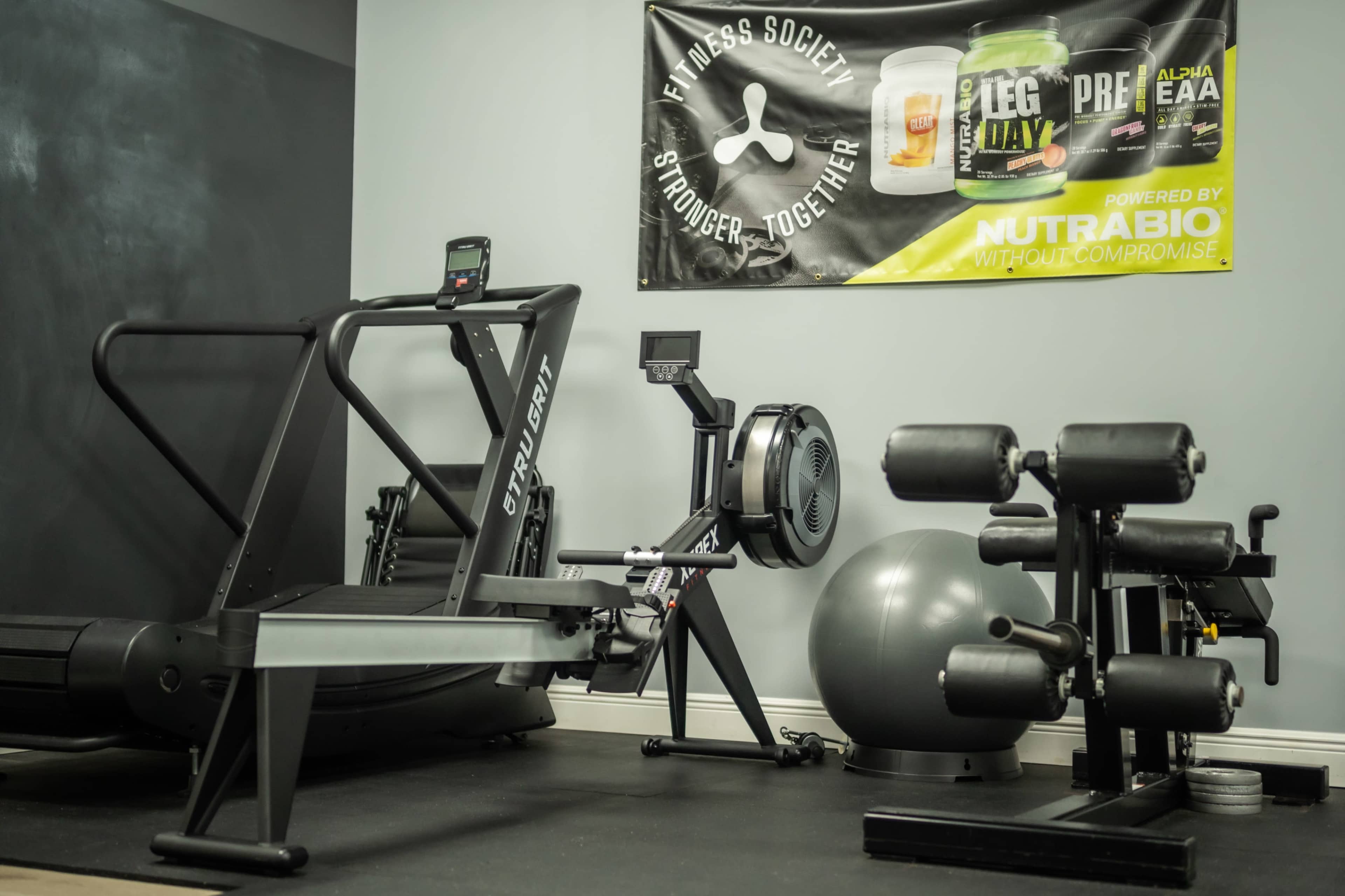 A home gym setup featuring a curved treadmill, a rowing machine, a stability ball, and a dumbbell rack under a promotional banner for a fitness brand.