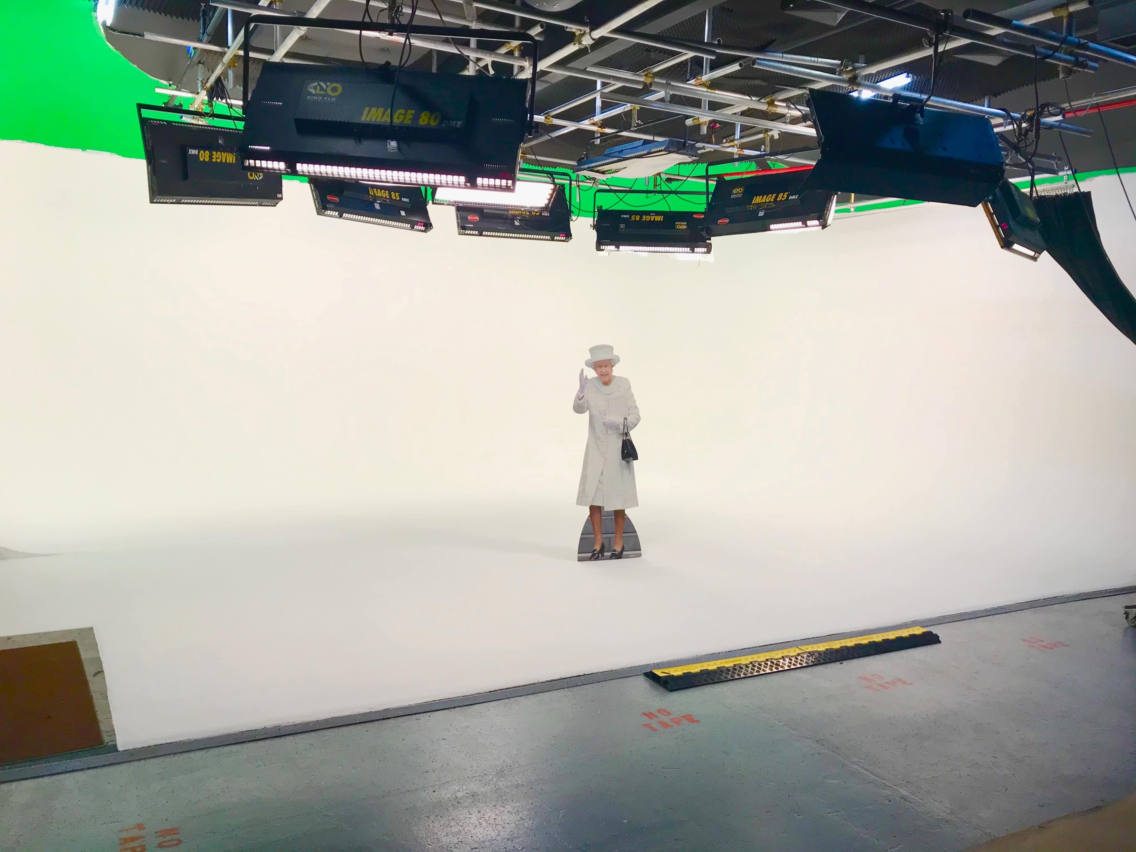 A lone figure dressed in a white outfit stands on a blank studio set, surrounded by bright lighting equipment and a green screen.