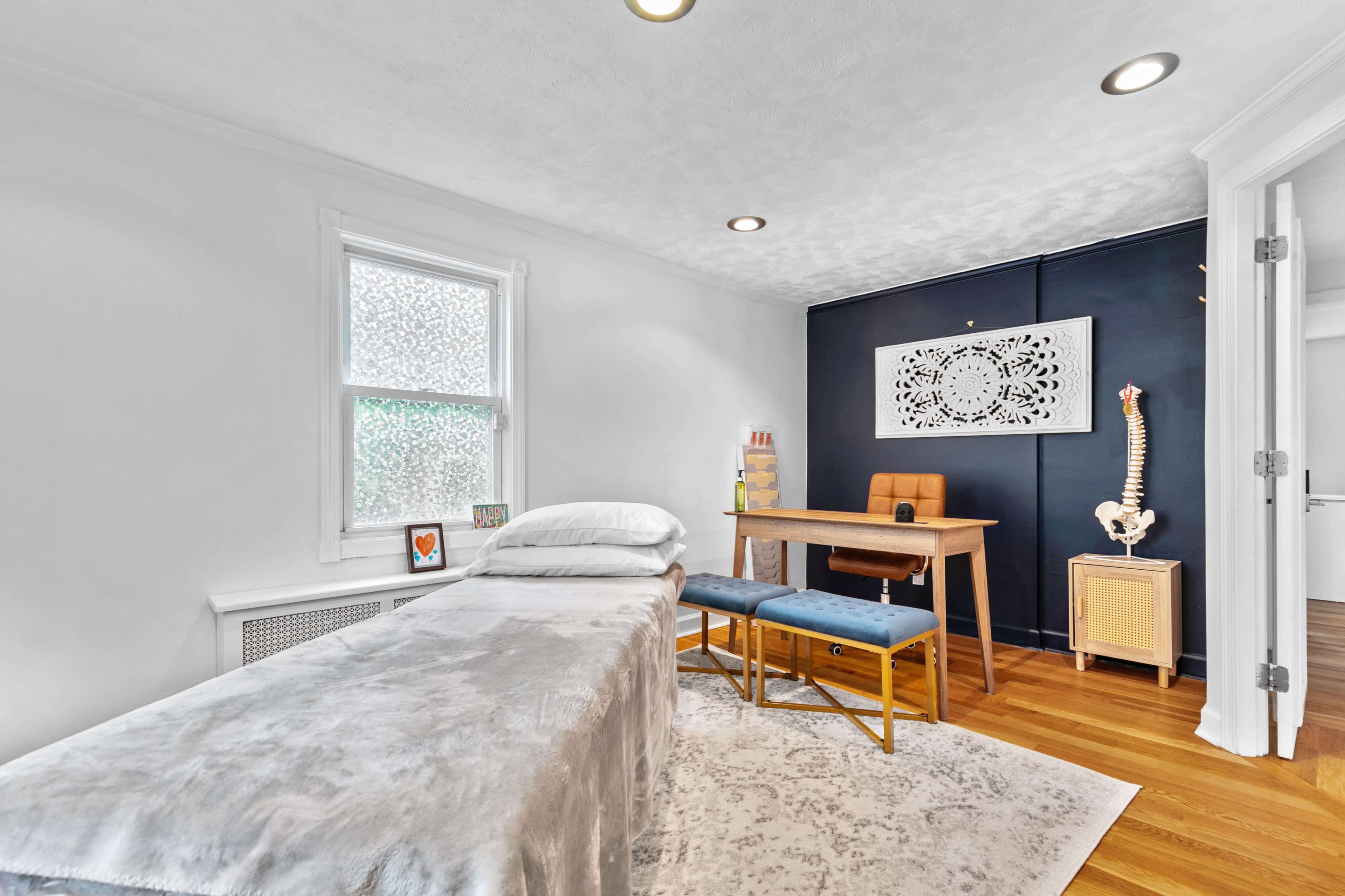 The room features a massage table covered with a gray blanket, a wooden desk with a chair, and a wall displaying an intricate white design, with natural light coming through a window.
