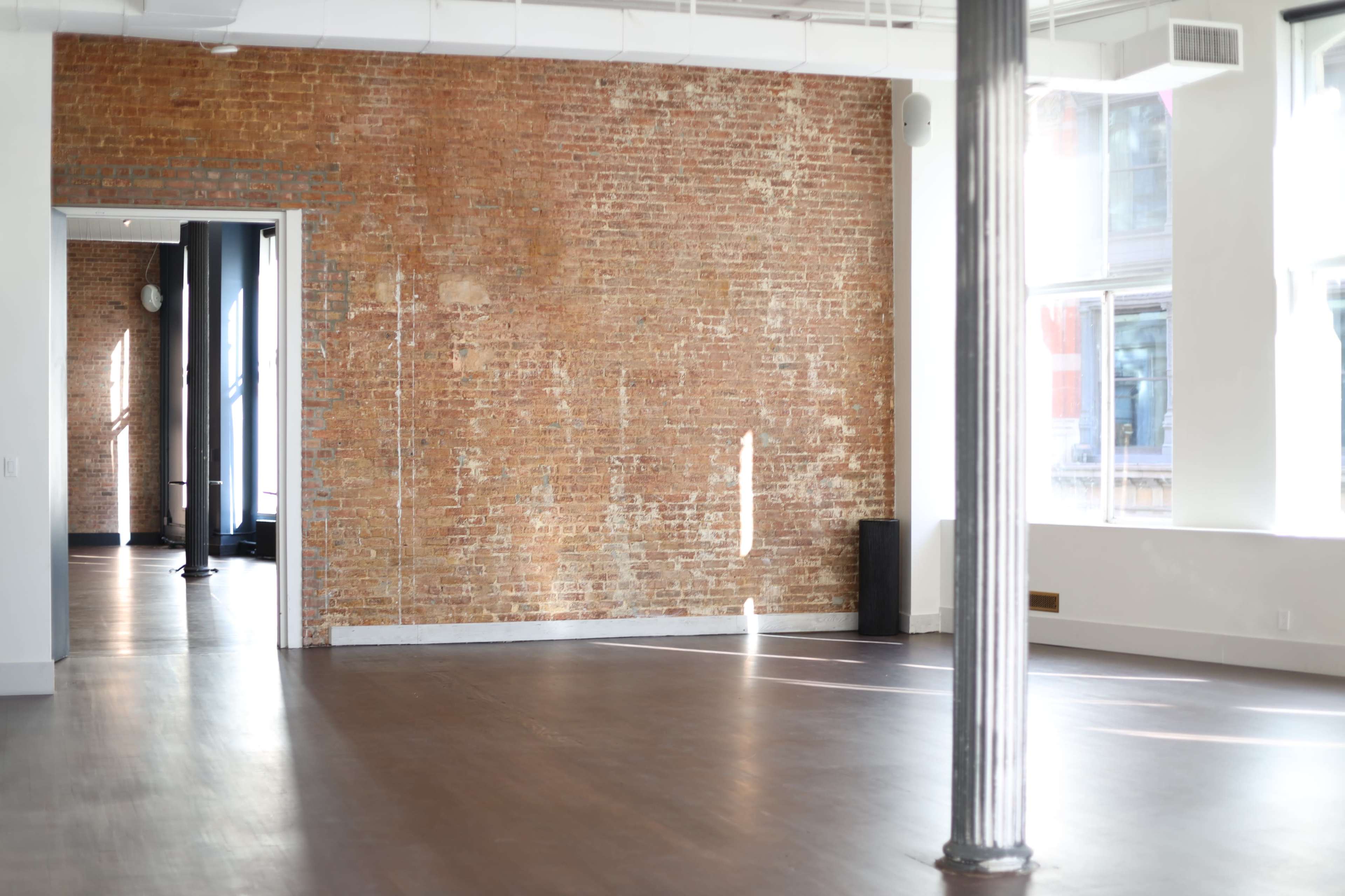 The image shows a spacious room with exposed brick walls, large windows, and polished wooden floors.