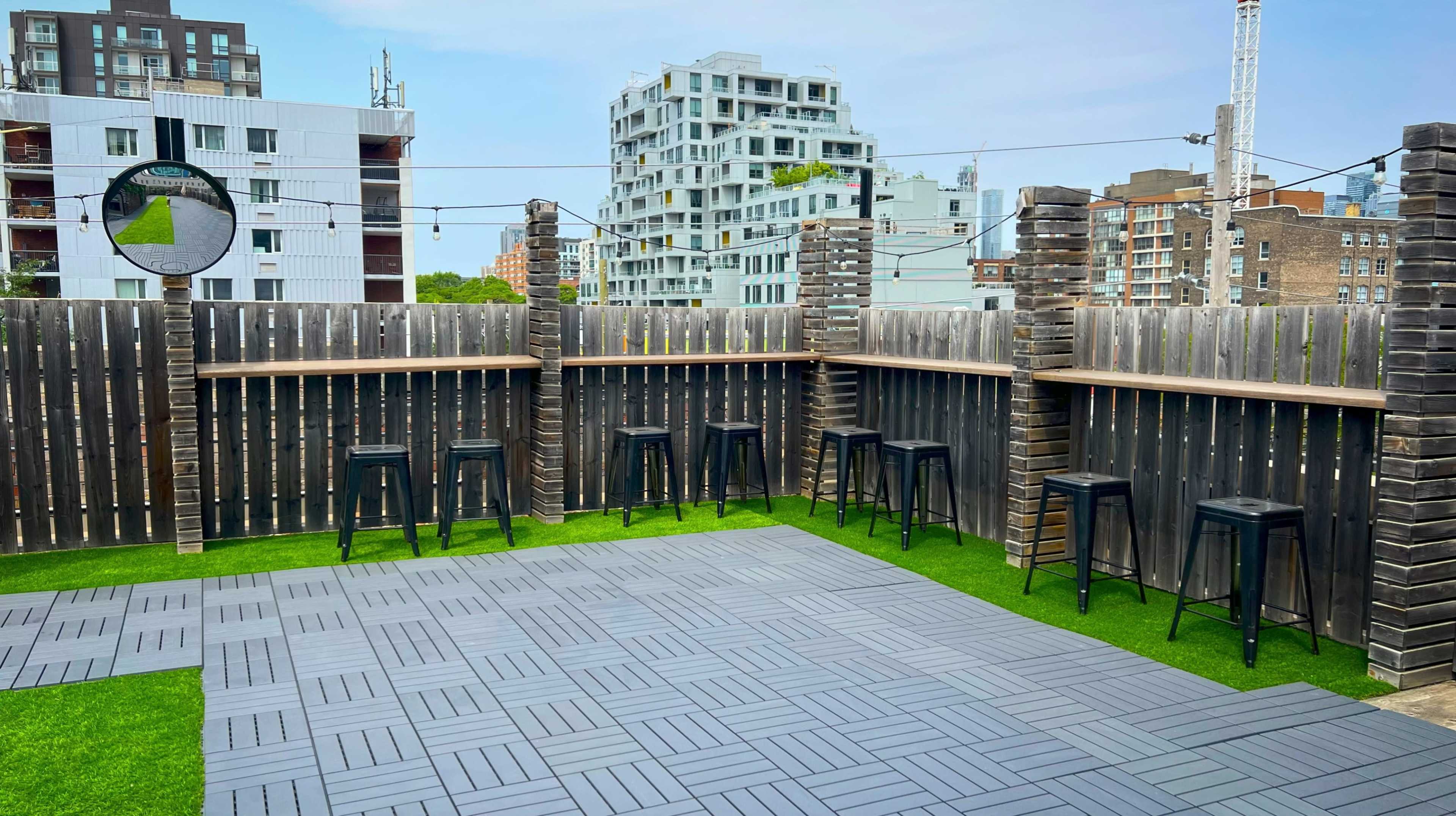 The image features a rooftop space with wooden fences, black bar stools lining a counter, and a modern city skyline in the background.