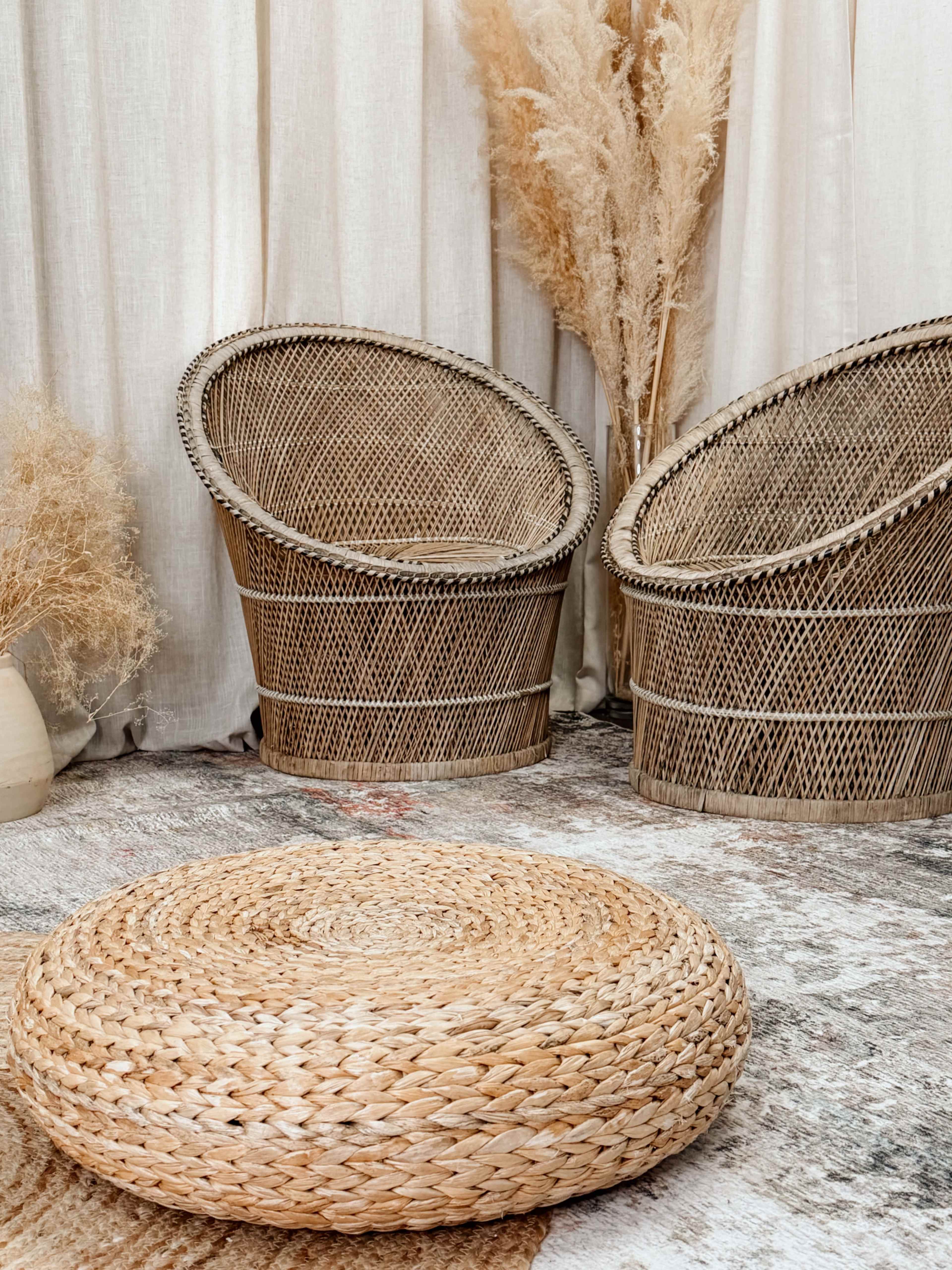 The image shows two wicker chairs and a round braided floor cushion in front of a light-colored fabric backdrop with dried plants.