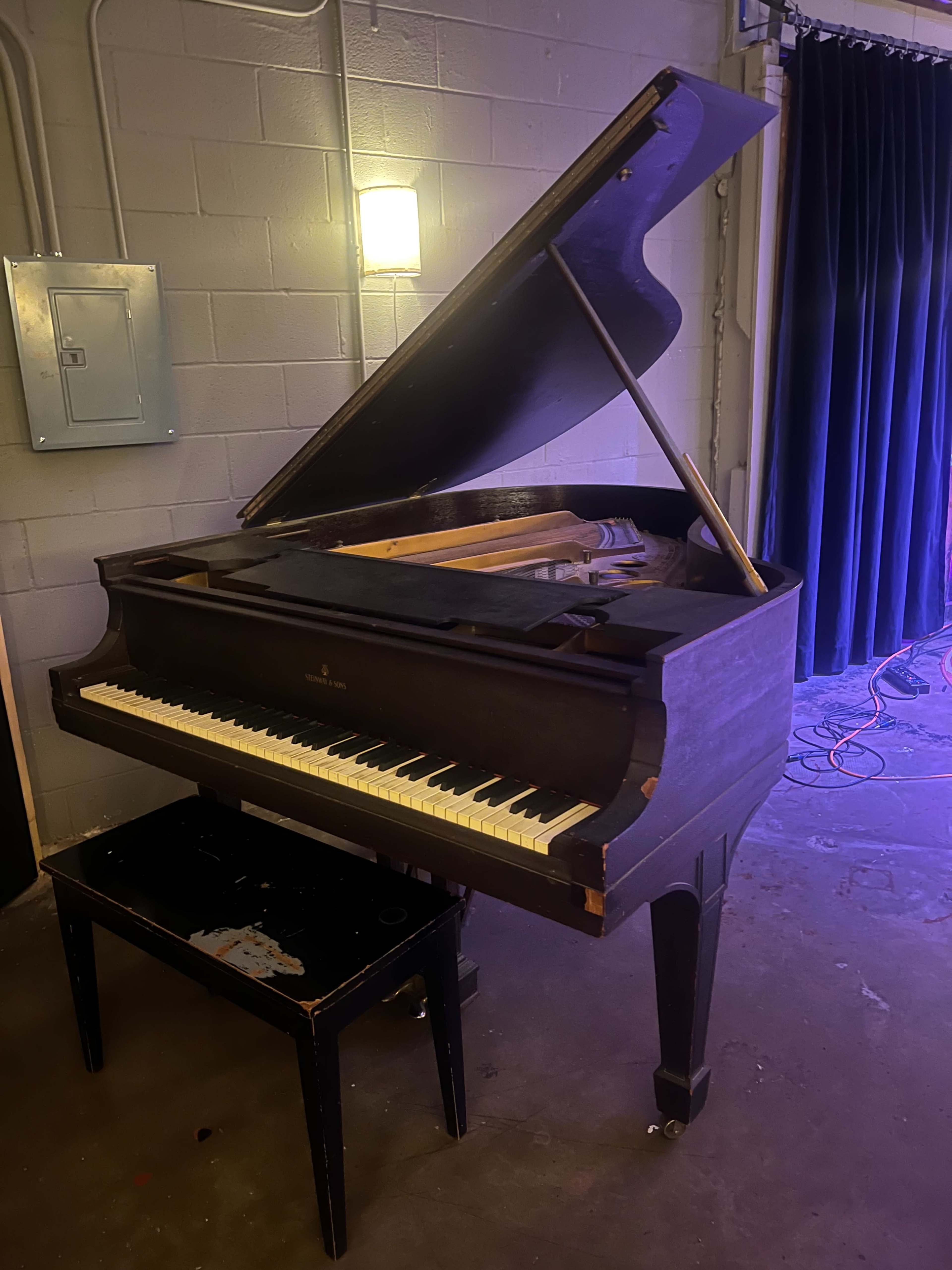 A grand piano is positioned on a black stool in a dimly lit room with a black curtain in the background.