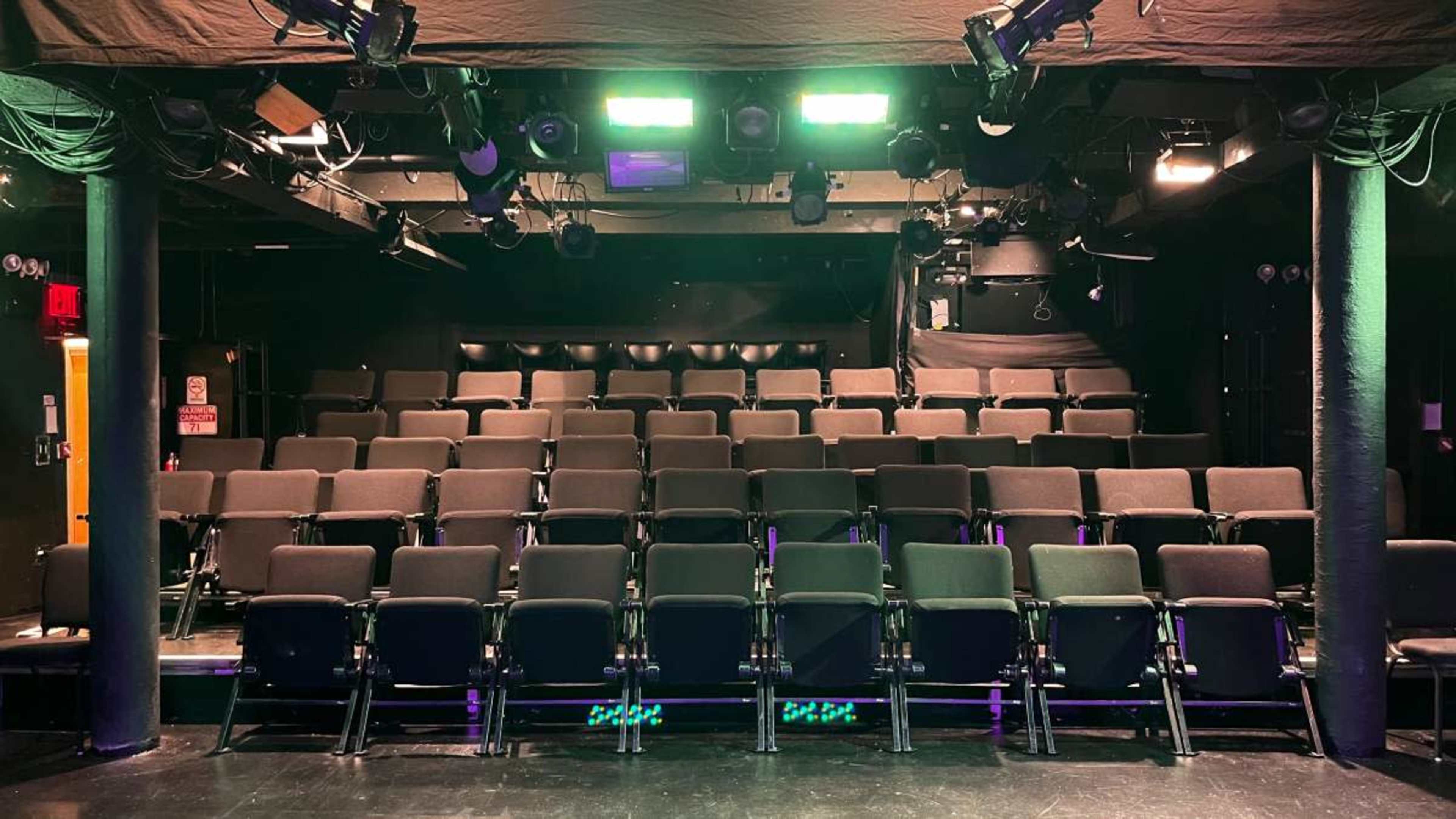 The image shows a darkened theater with rows of black folding chairs arranged in an ascending layout.