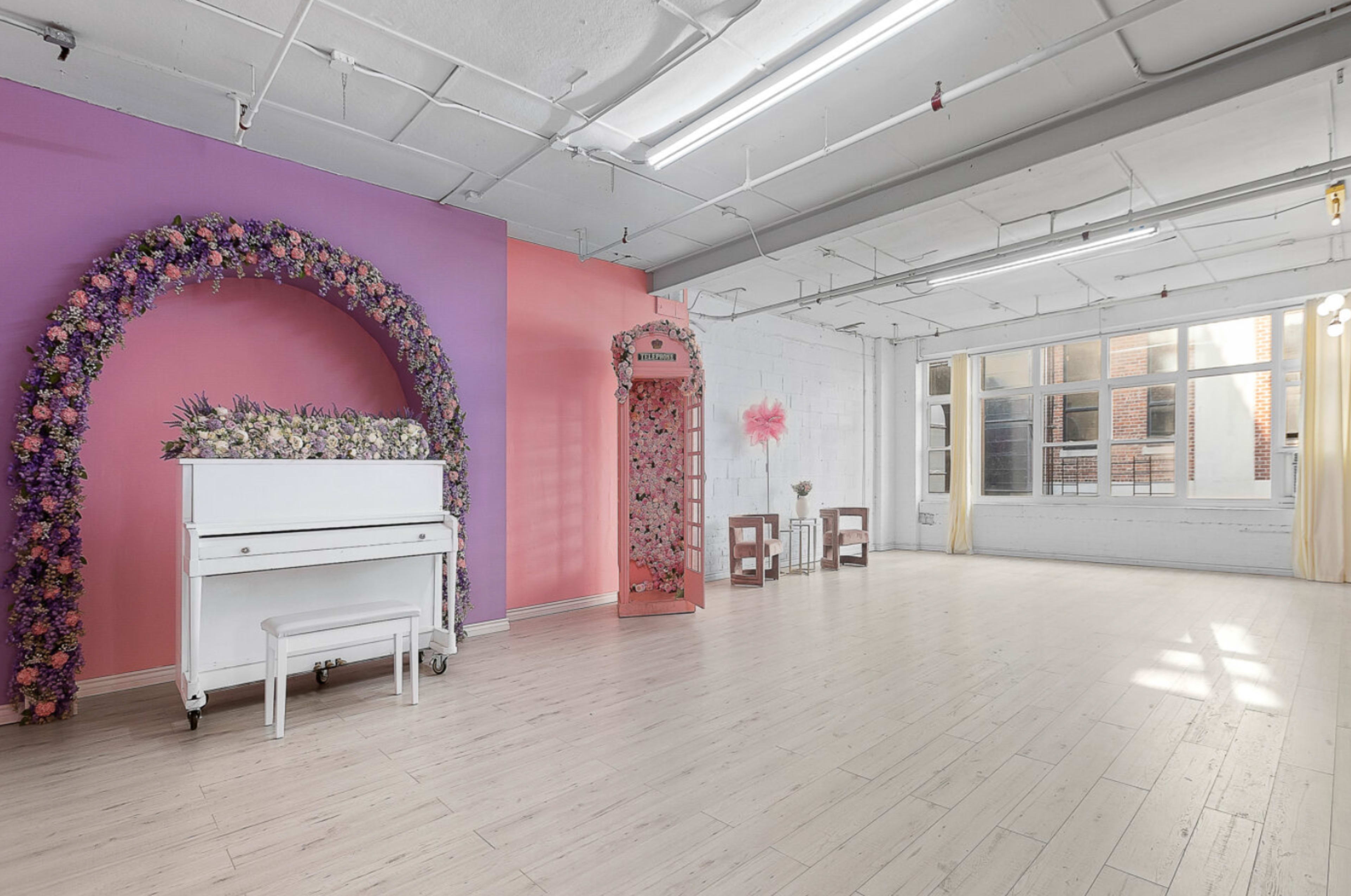 A brightly lit, spacious room featuring a white piano under a floral arch and a pink corner structure adorned with flowers.