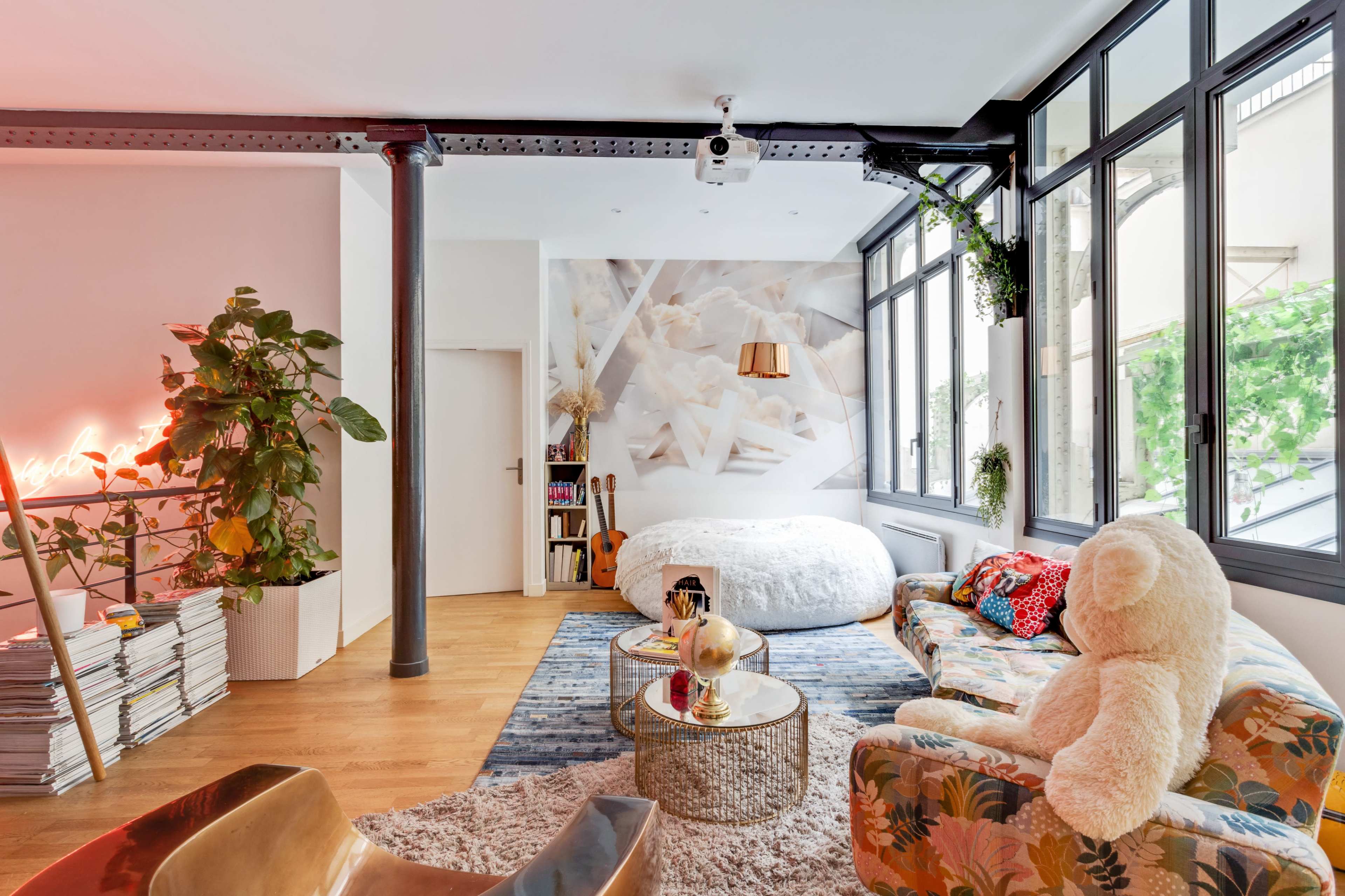 The image shows a stylish, modern studio apartment featuring a round bed, a large window area with greenery outside, a cozy seating arrangement, and decorative elements like a giant teddy bear and a neon sign.