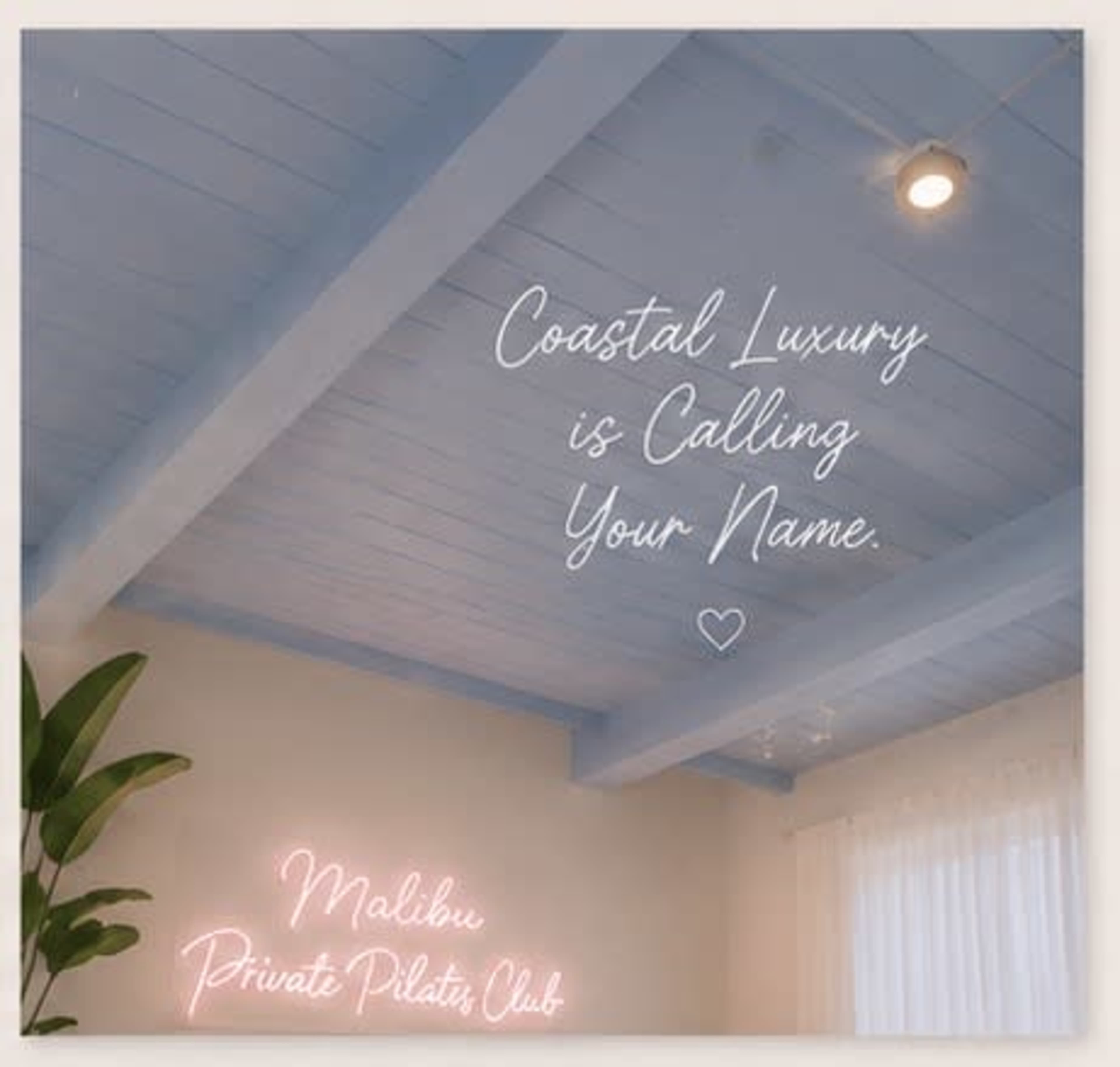 The image features a room with light blue wooden ceiling beams and a wall adorned with decorative text that reads "Coastal Luxury is Calling Your Name" alongside a neon sign that says "Malibu Private Pilates Club."
